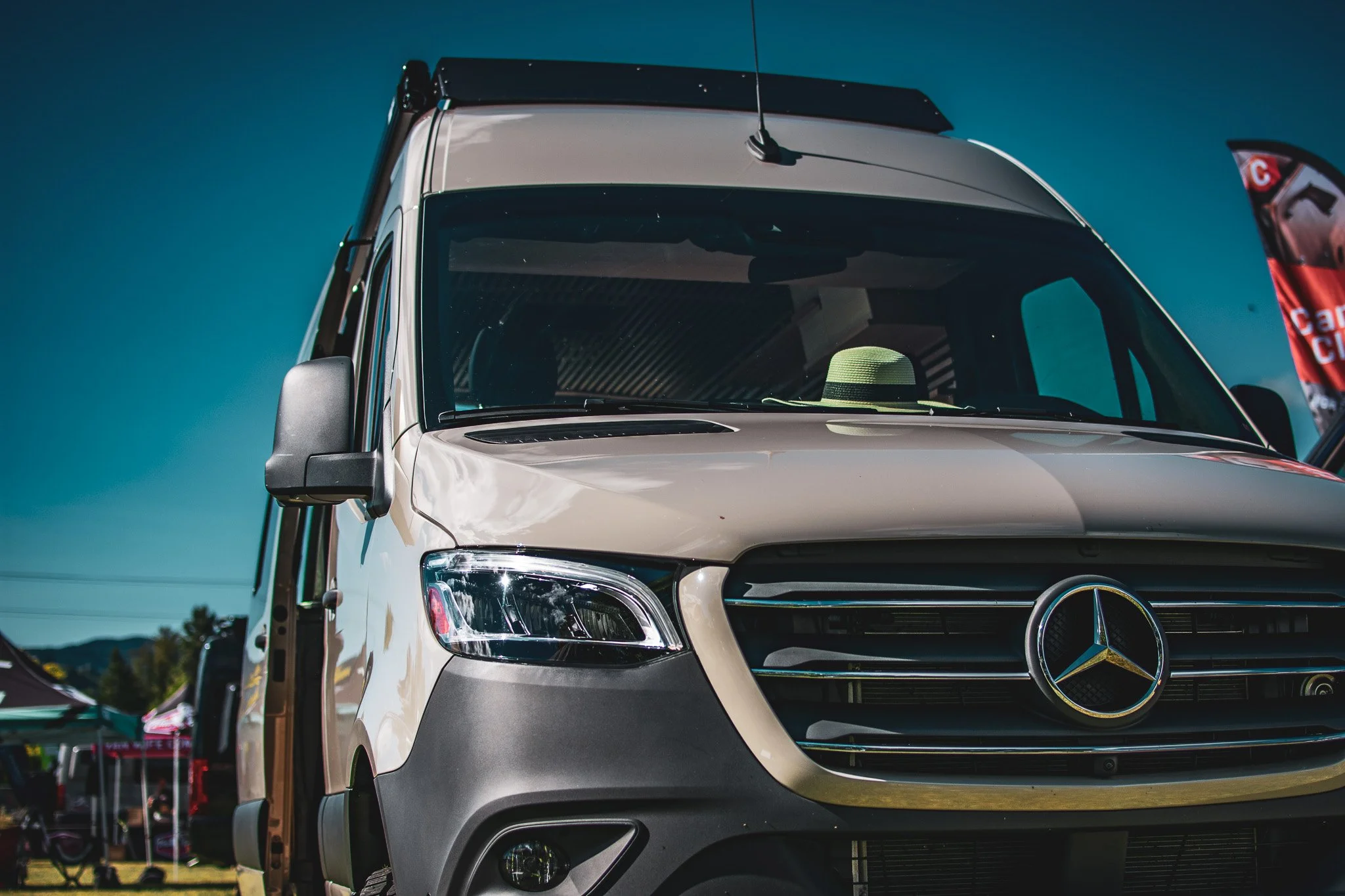 Front view of a white Mercedes-Benz van with a prominent Mercedes logo on the grille, parked outdoors on a sunny day, with tents and flags in the background.