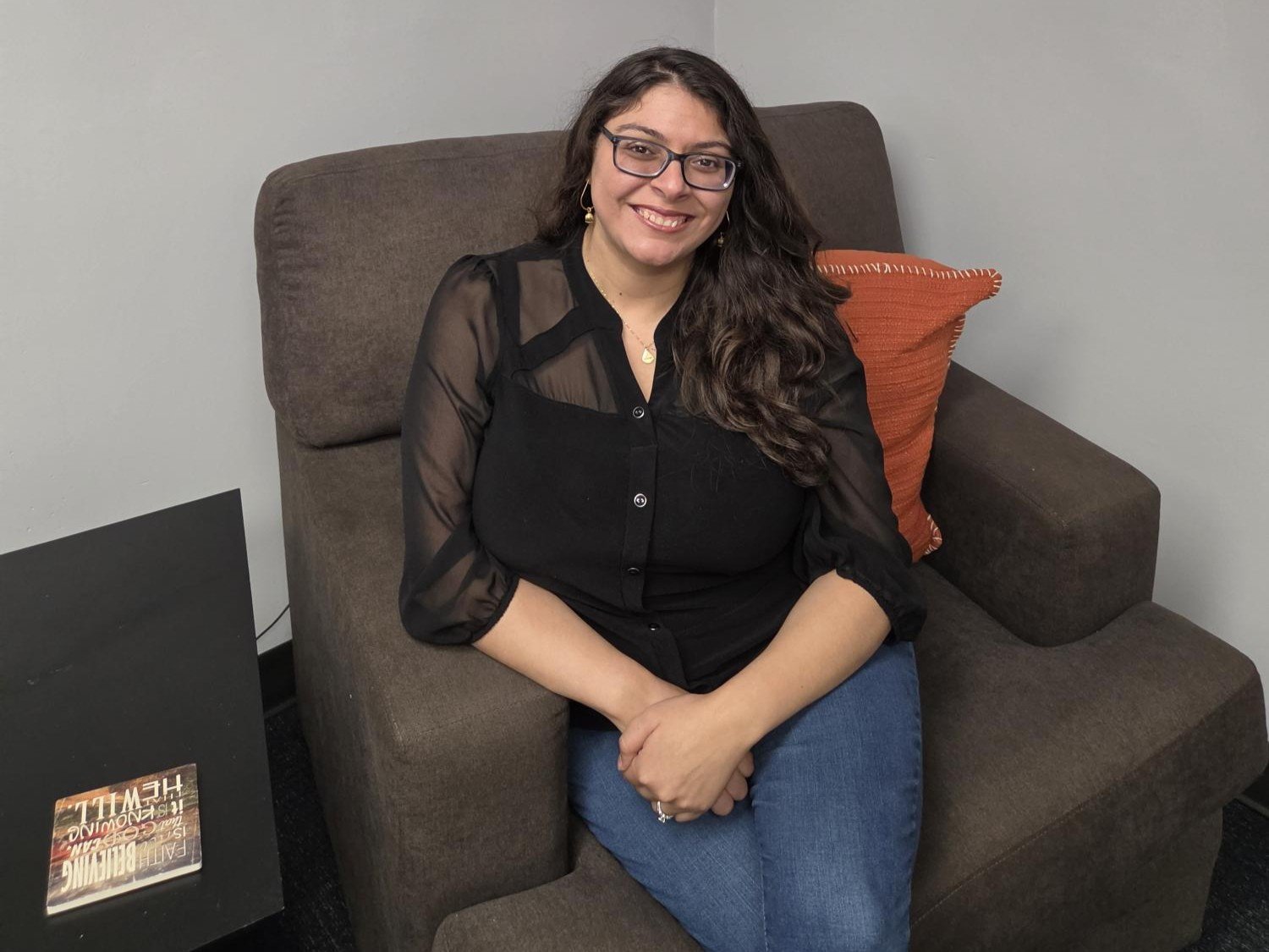 A woman with long dark hair, glasses, and earrings sitting on a brown couch in a room with beige walls. She is smiling, wearing a black blouse with sheer sleeves and blue jeans. There is an orange pillow behind her and a black table with a book on it nearby.