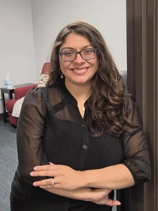 A woman with long, curly brown hair and glasses smiling at the camera, sitting in a room with gray walls, red couch, and tissue box on a side table.