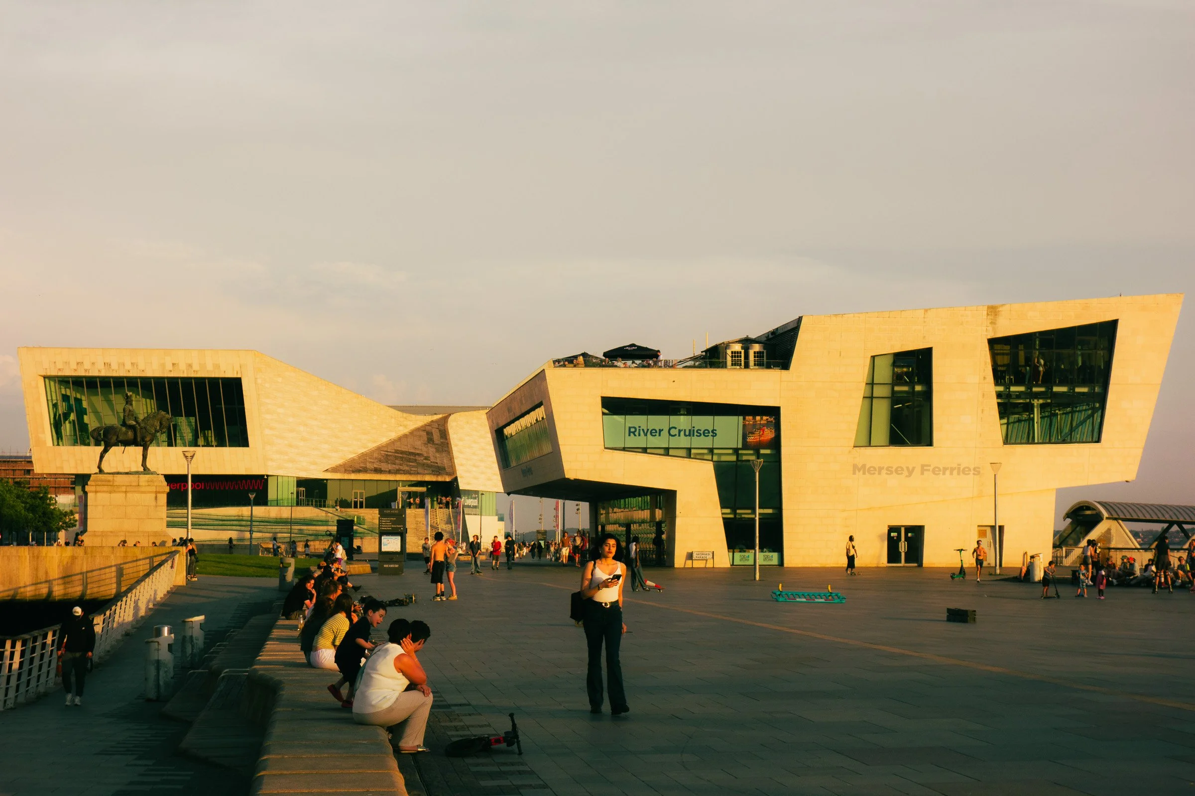 Modern building with large glass windows, a statue of a horse and rider, and a sign for River Cruises, with people walking and sitting in the foreground under a cloudy sky.