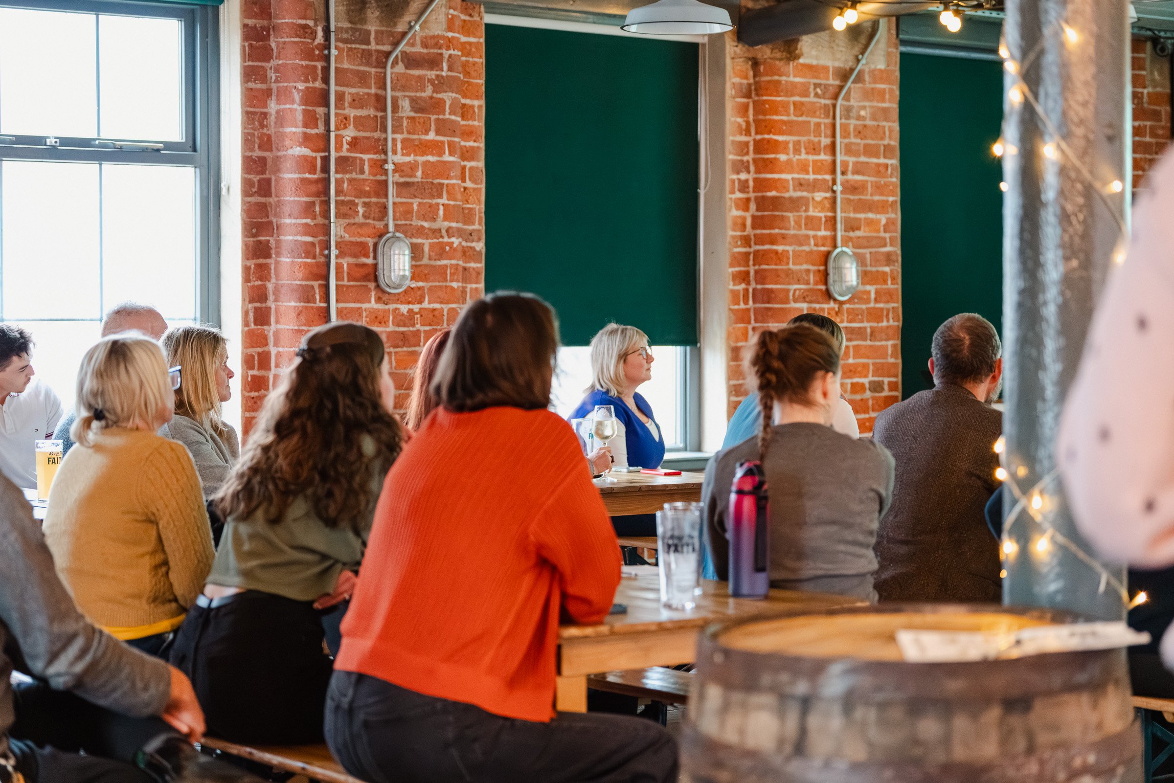 A group of people gathered indoors, sitting at tables and listening attentively. The setting features with exposed brick walls, large windows allowing natural light, and some string lights. Some individuals are holding drinks, and the atmosphere appe