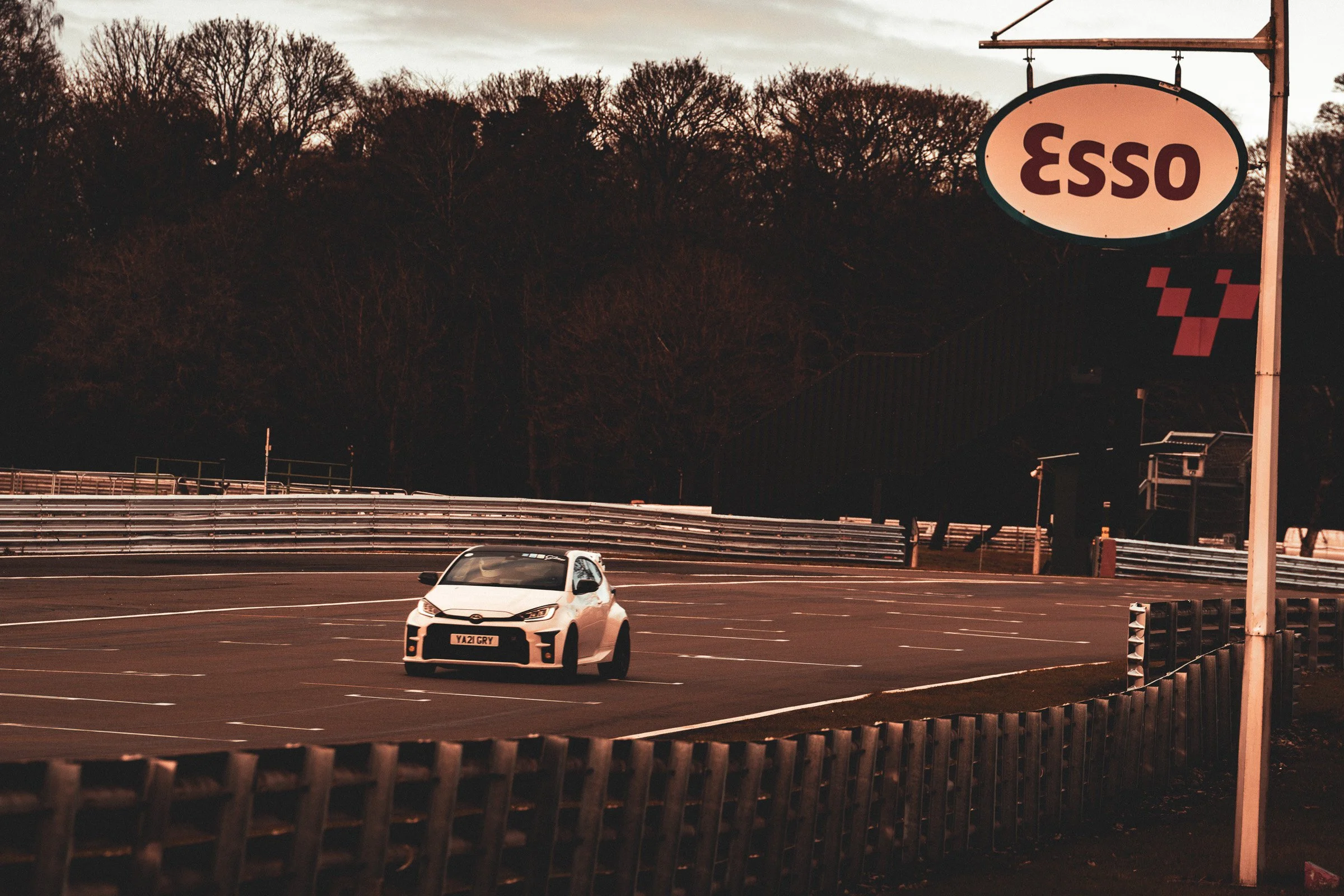 An empty parking lot with a white car and an Esso sign, surrounded by a wooden fence and trees in the background at dusk.