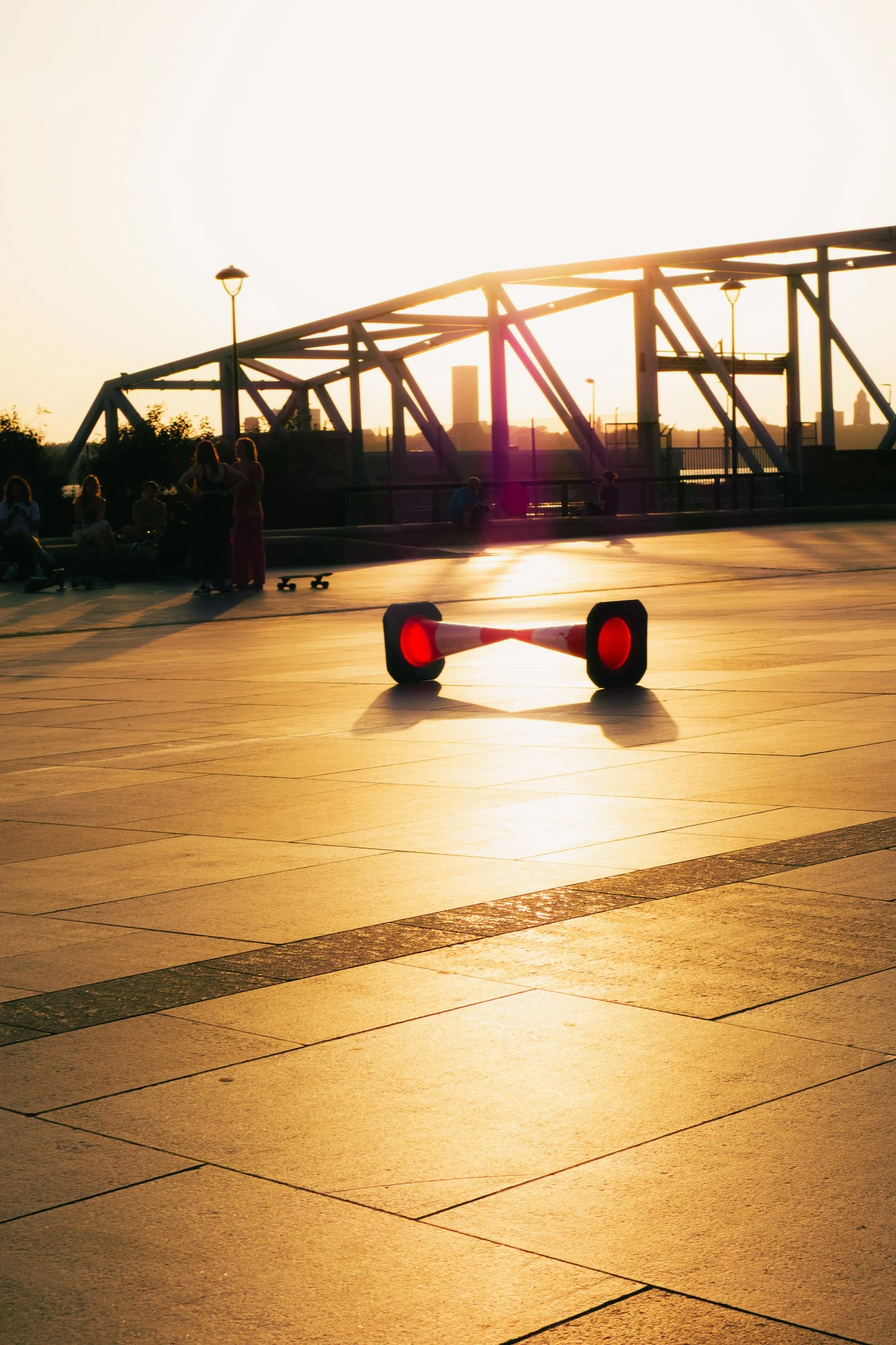People sitting and standing on a terrace with skateboards during sunset, with a bridge and city skyline in the background.