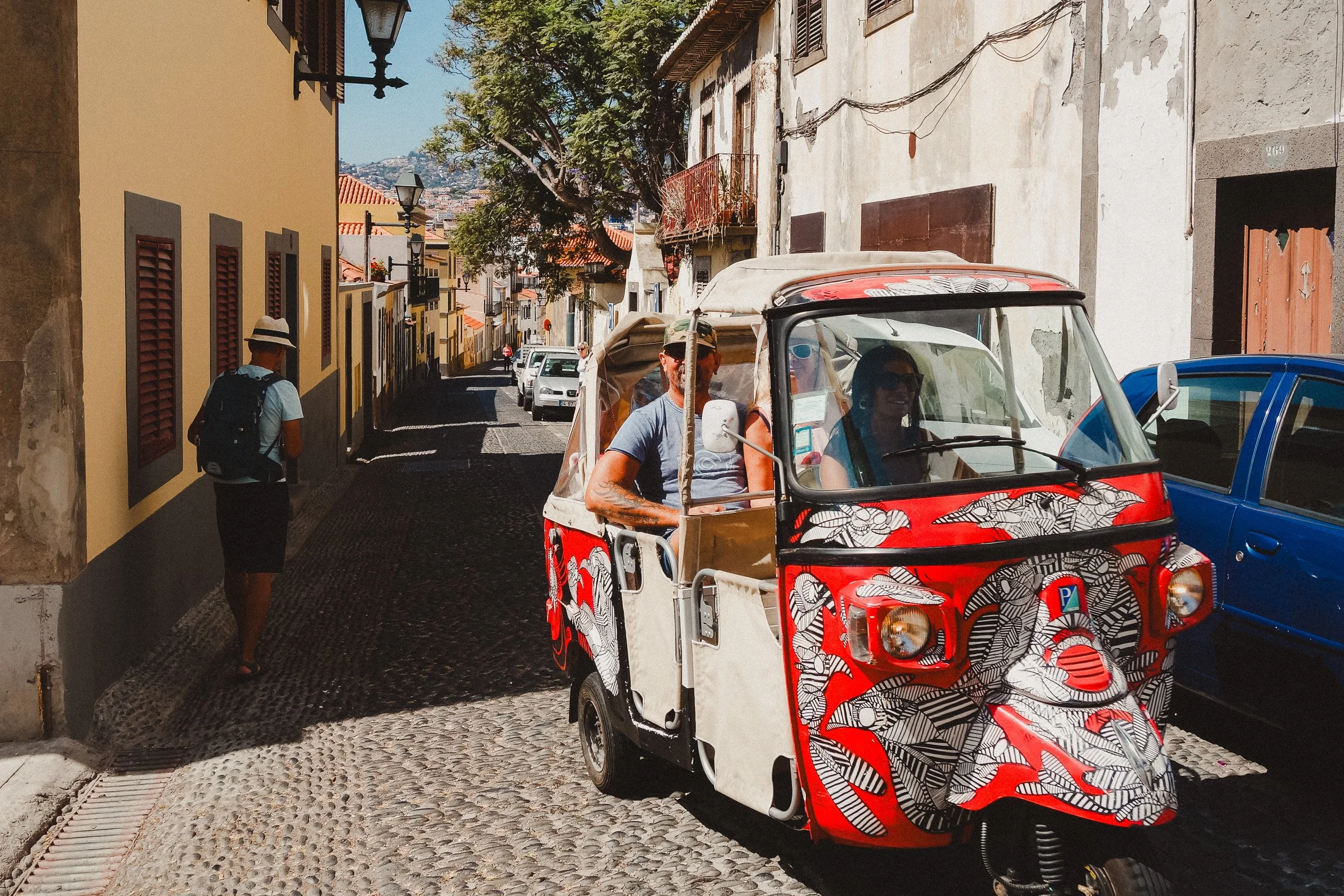 A street scene with colorful buildings, a tree, and a small red and white tuk-tuk vehicle with a patterned design. Parked cars line the narrow street in Funchal, Madeira. The sky is clear and sunny.