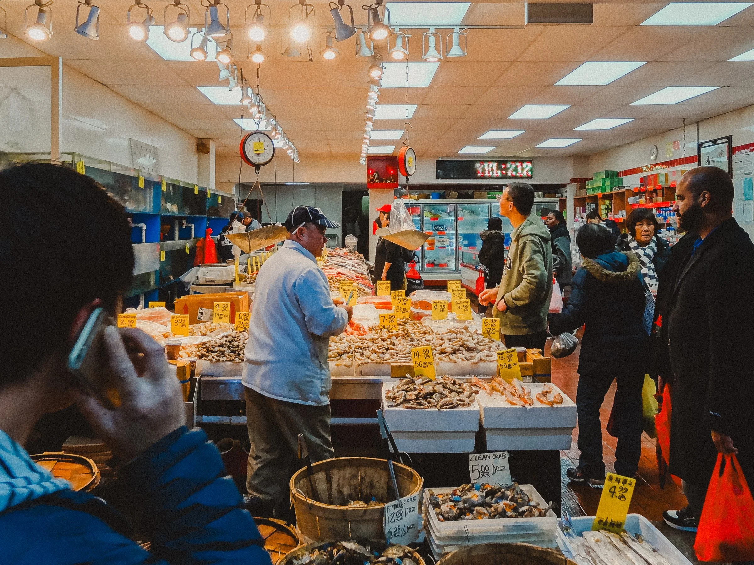 People shopping for seafood at a fish market, with various fish displayed on ice and yellow price signs, and overhead lights illuminating the area.
