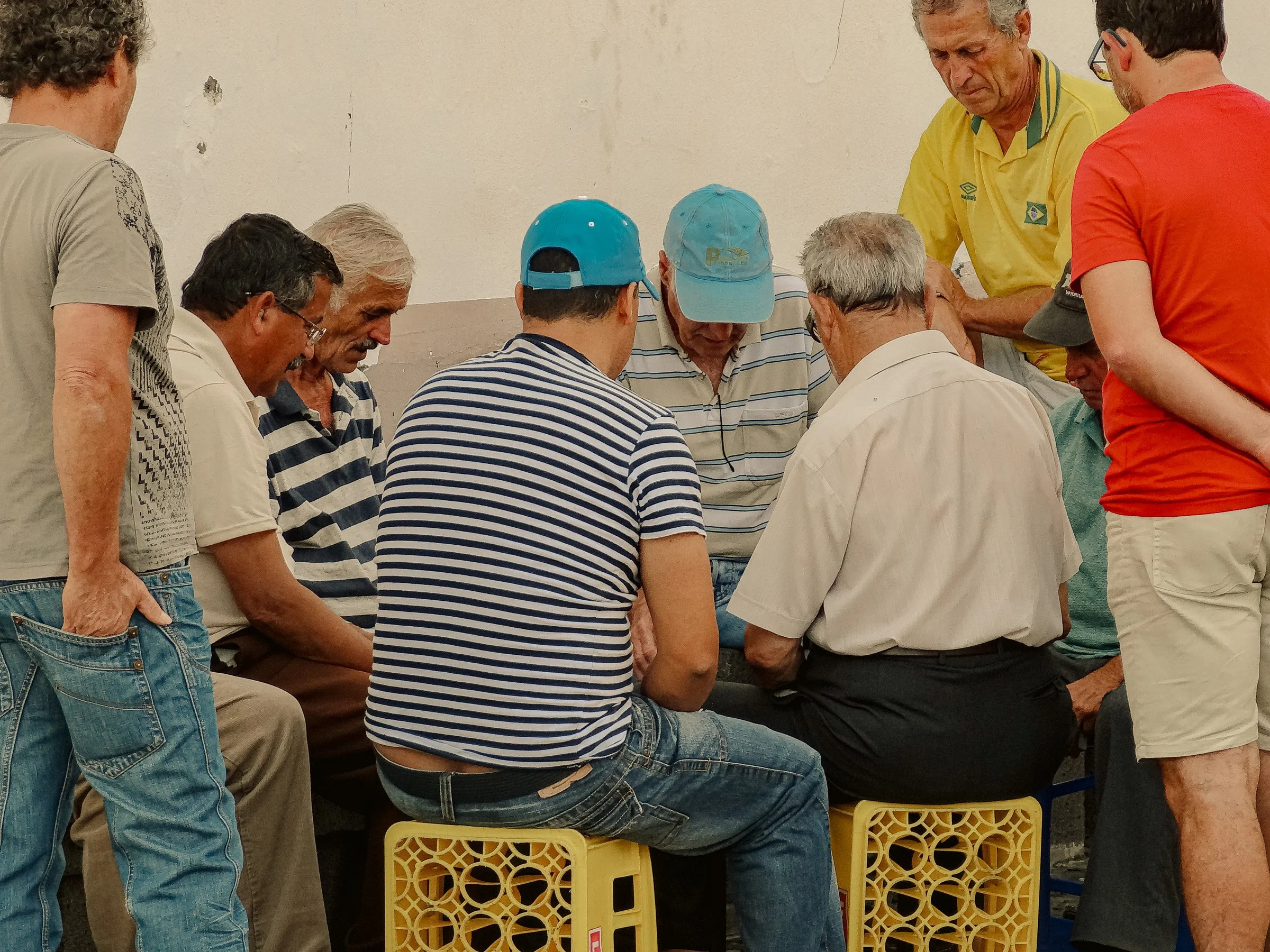 A group of men gathered closely together, some standing and some sitting, engaged in a game of cards, with stools and a plain wall in the background.