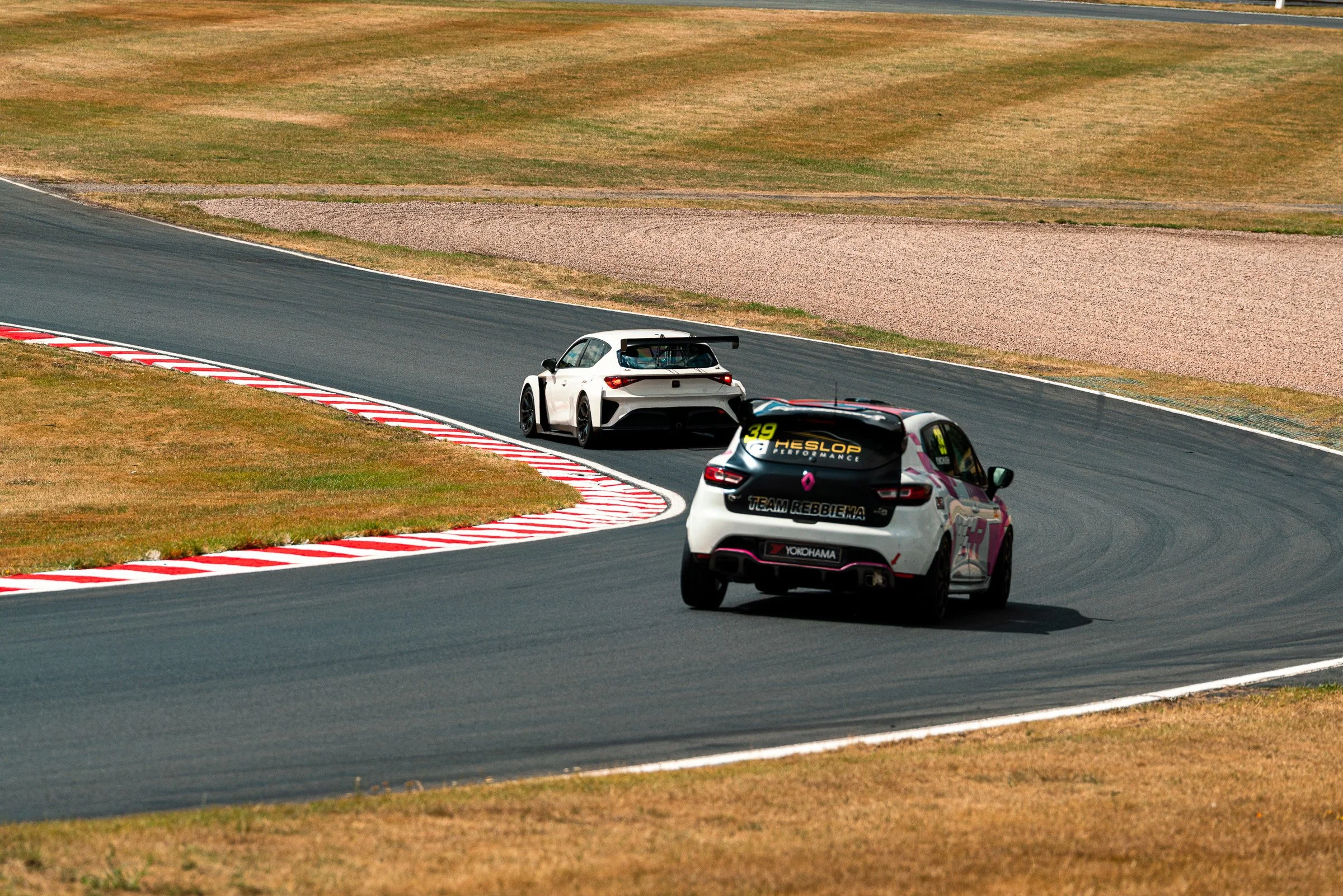 Two race cars on a racetrack, navigating a curve with red and white painted curb markings, surrounded by dry grass.