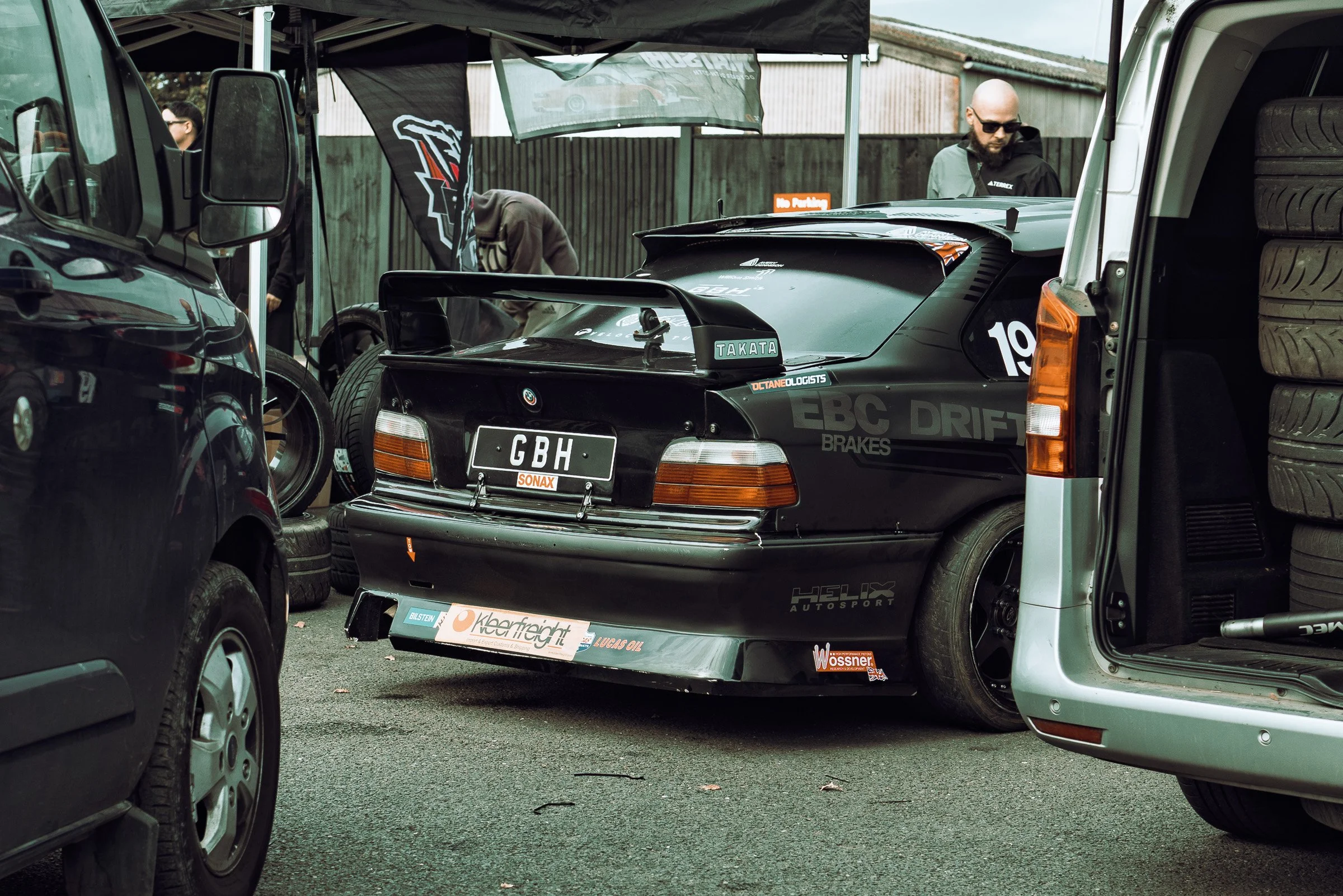 A black race car with a large rear wing and sponsor stickers, parked between several cars and a stack of tires at a racing event.