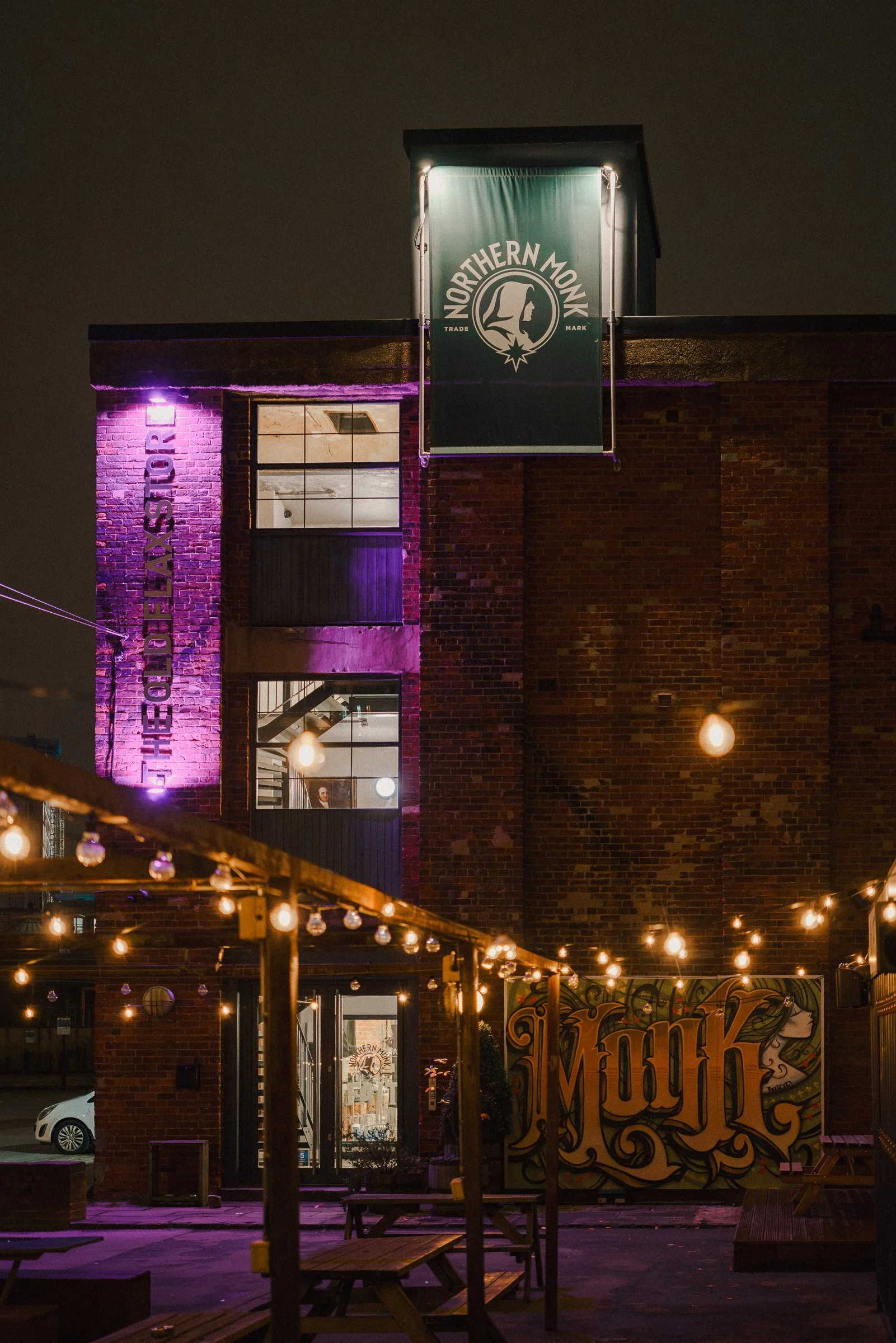 Night view of a brick building with illuminated signs and string lights hanging in an outdoor patio area. A green banner with 'Northern Monk' logo is visible. The area appears lively and inviting with outdoor seating.