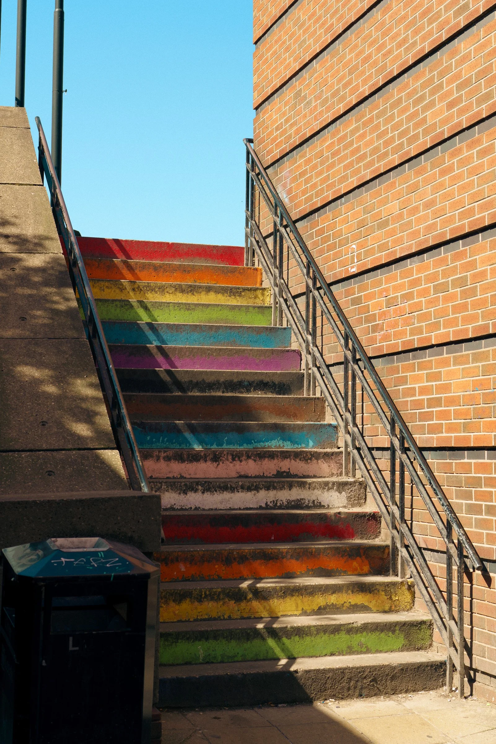 Rainbow-colored painted stairs leading up along a brick building, with a metal railing on the right, and a trash bin at the bottom left corner.
