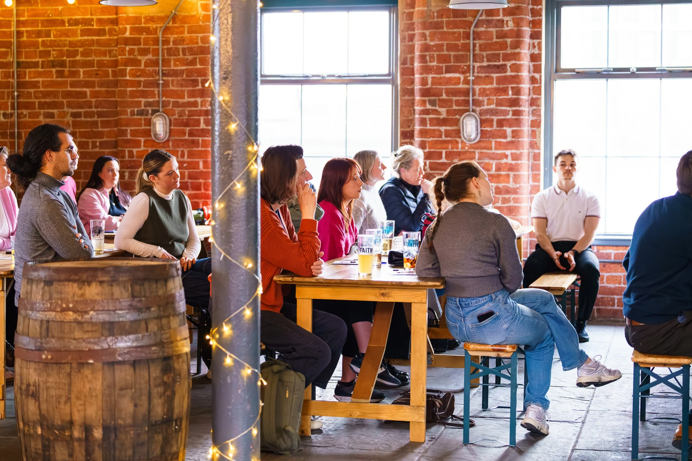 A group of diverse people sitting at picnic tables in a rustic indoor space with exposed brick walls and large windows, attentively listening to a speaker or presentation.