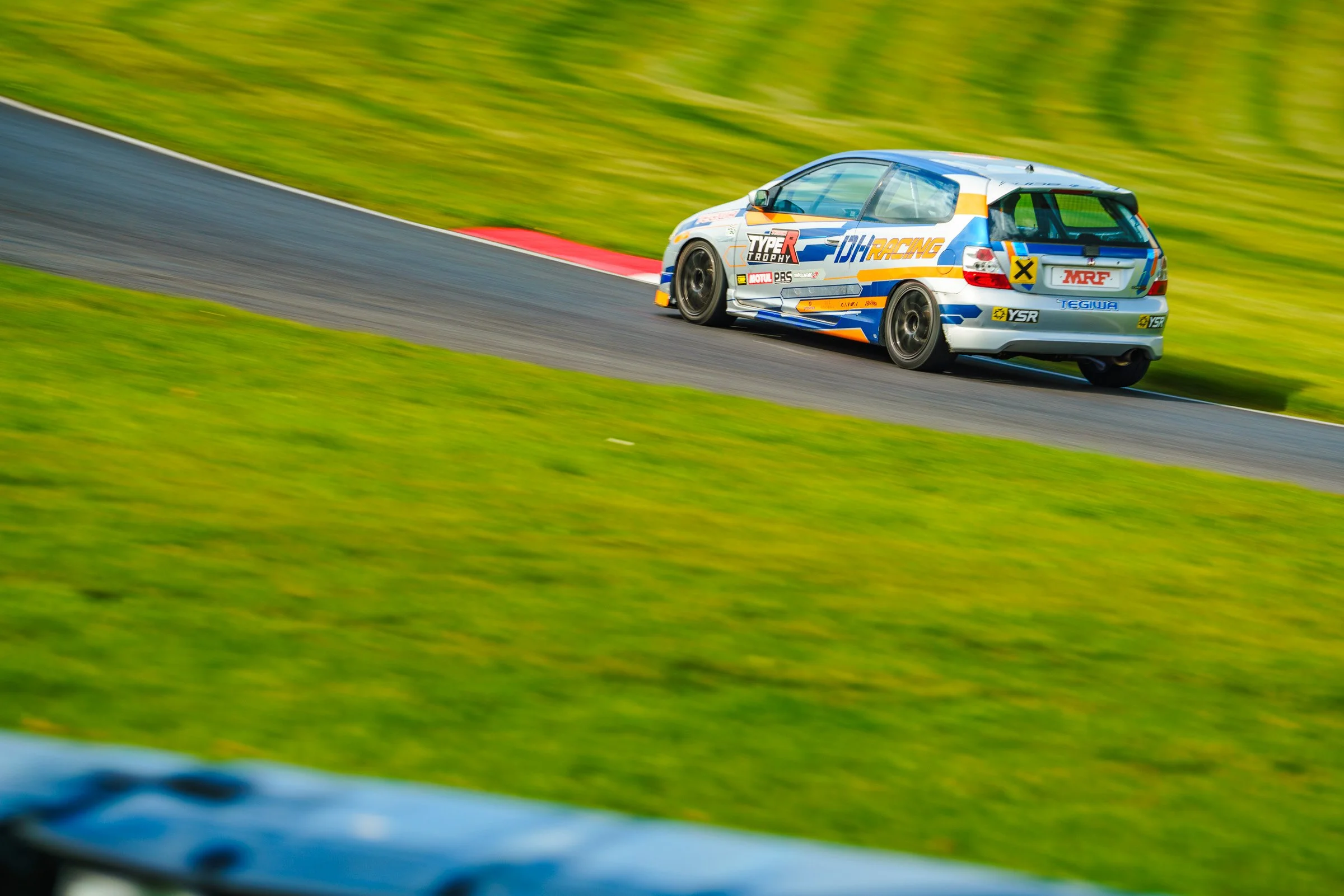 A white race car with blue and orange markings speeding on a racetrack with green grass on both sides.
