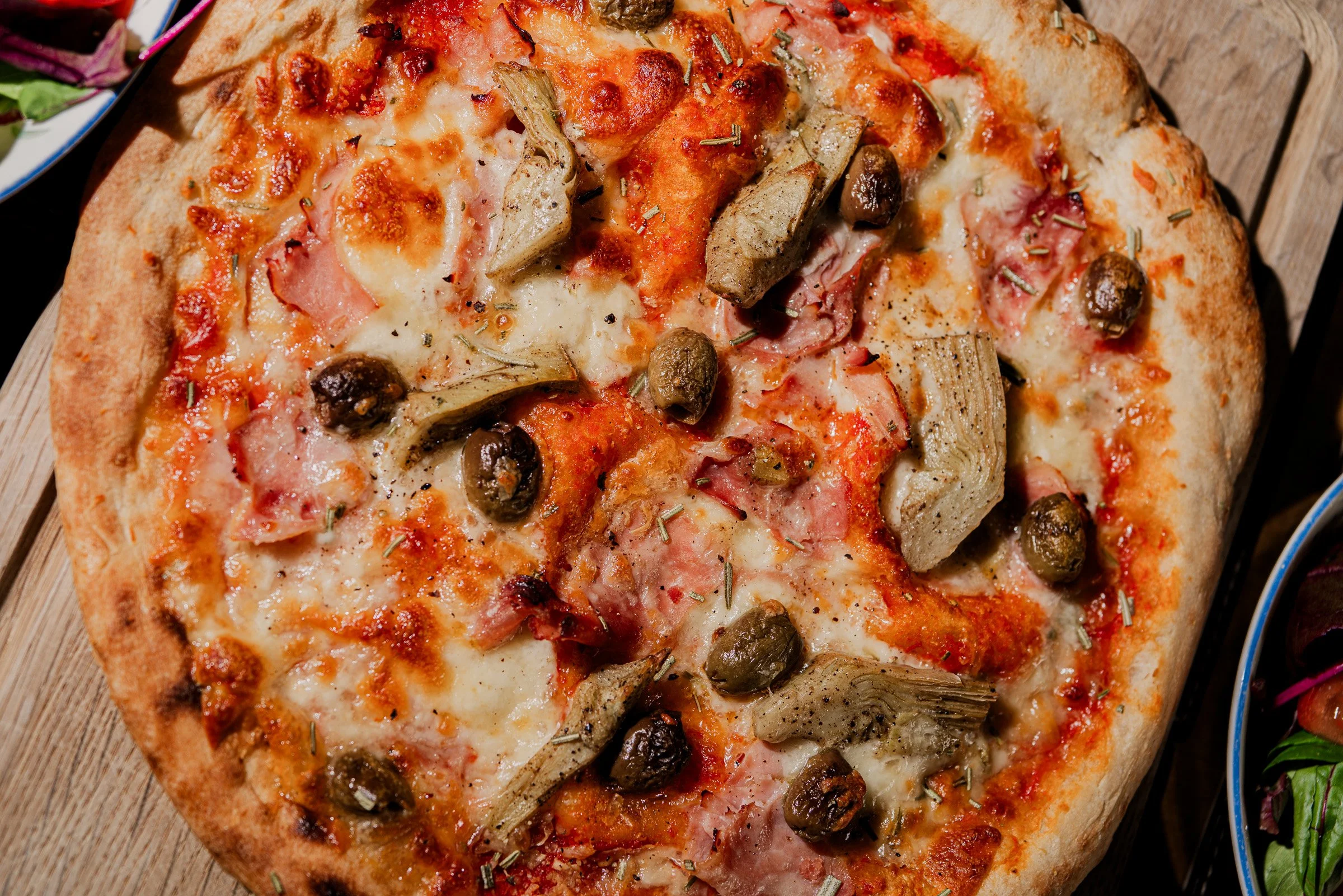 Close-up of a freshly baked pizza with cheese, black olives, artichoke hearts, and ham on a wooden surface.