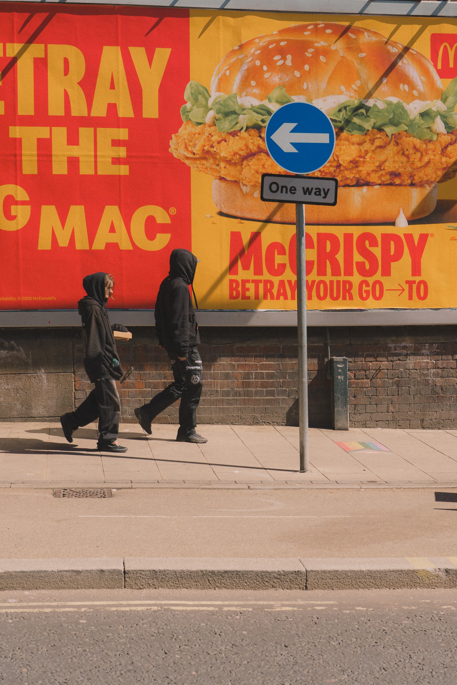 A street scene with two people in hoodies walking past a large McDonald's advertisement for a chicken sandwich. The advertisement features a burger with fried chicken, lettuce, and sesame seed bun. There is a street sign indicating a one-way street w