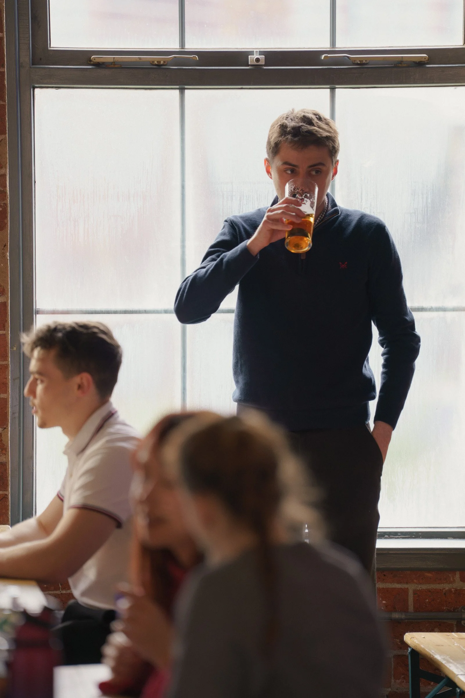 A young man in a dark sweater drinking beer from a glass in a casual indoor setting with other people around.