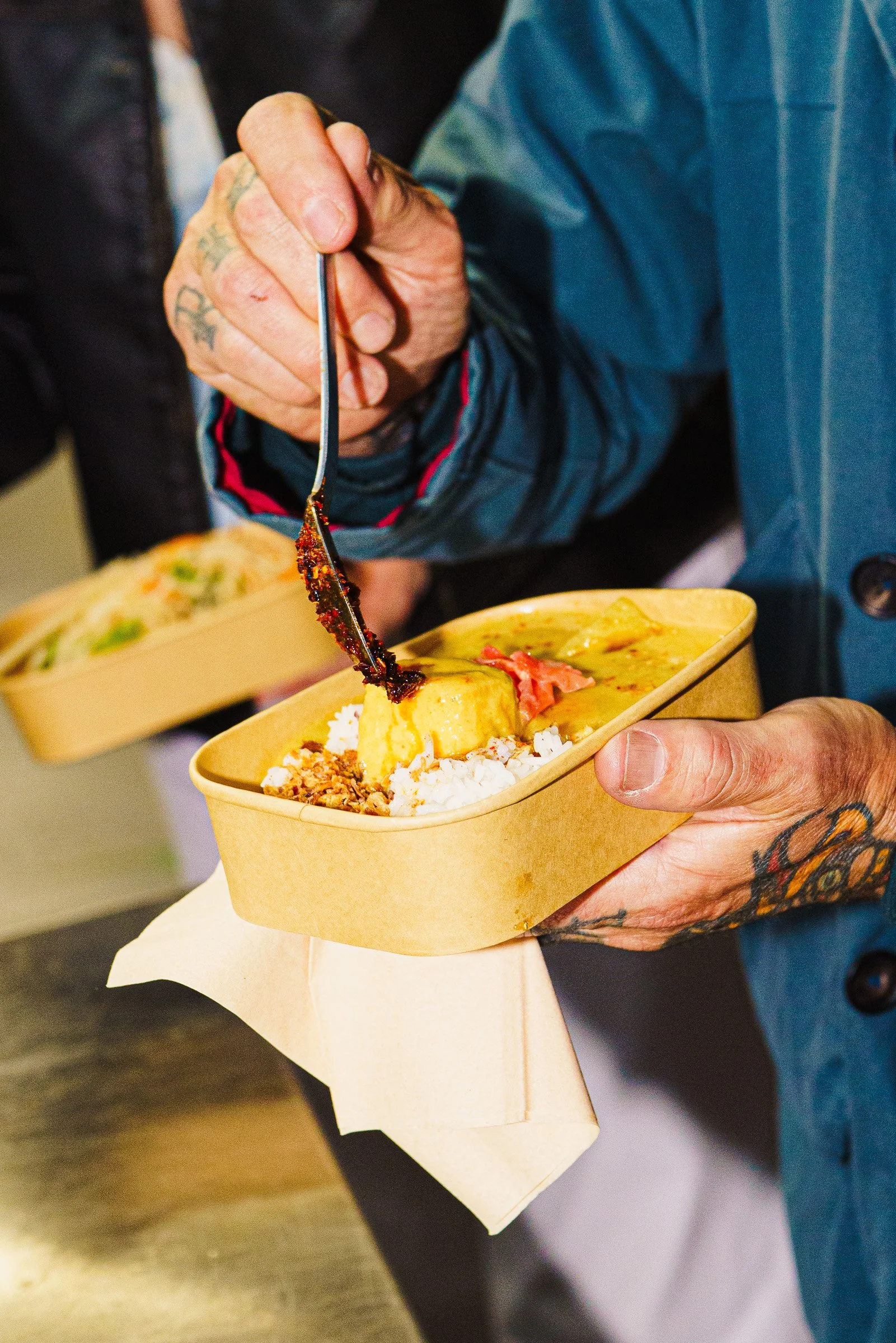 Close-up of a hand with tattoos holding a biodegradable food container filled with rice, a vegan Thai yellow curry, and toppings, as someone squeezes spicy chili sauce.