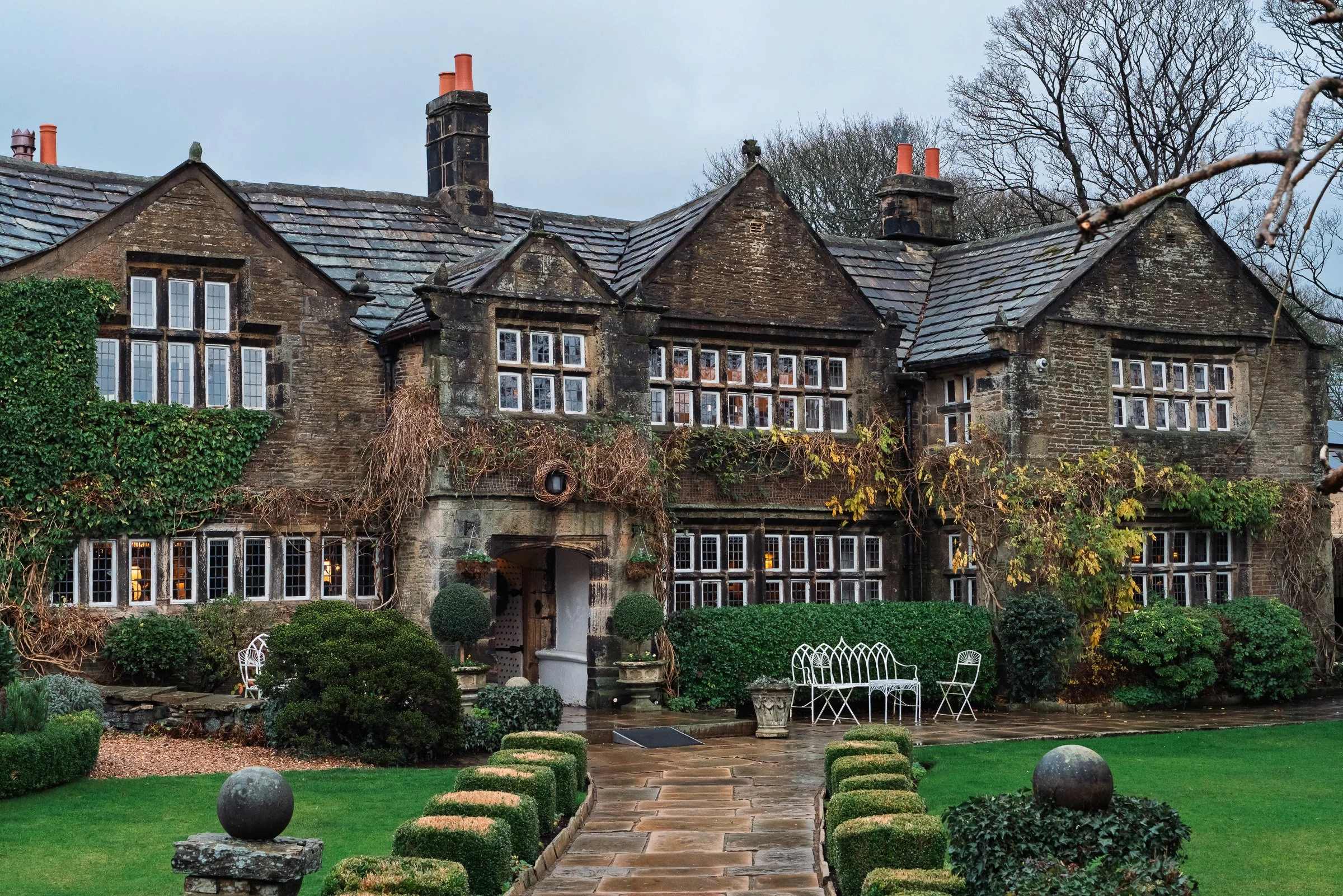 Historic stone mansion with multiple chimneys, small windows, and ivy-covered walls, surrounded by a neatly maintained garden with manicured bushes, small trees, and outdoor seating, on a cloudy day.
