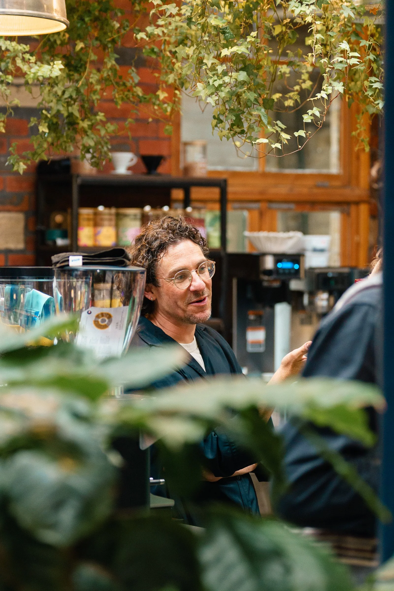 A woman with curly hair and glasses talking to someone in a cozy, rustic indoor space with plants hanging from the ceiling, a brick wall, and shelves with containers in the background.