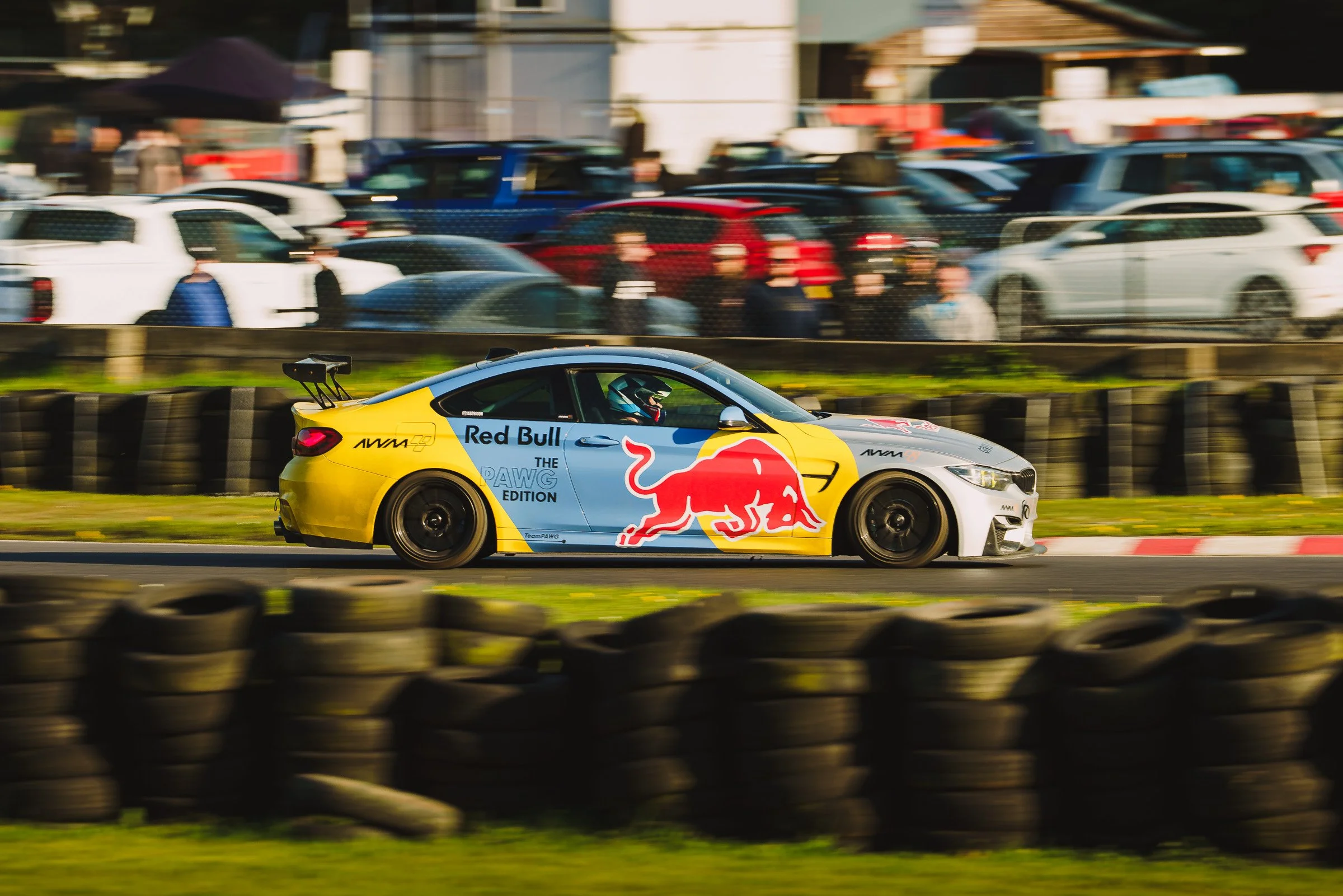 A BMW M4 race car with Red Bull branding driving on a racetrack, with blurred spectators and cars in the background.
