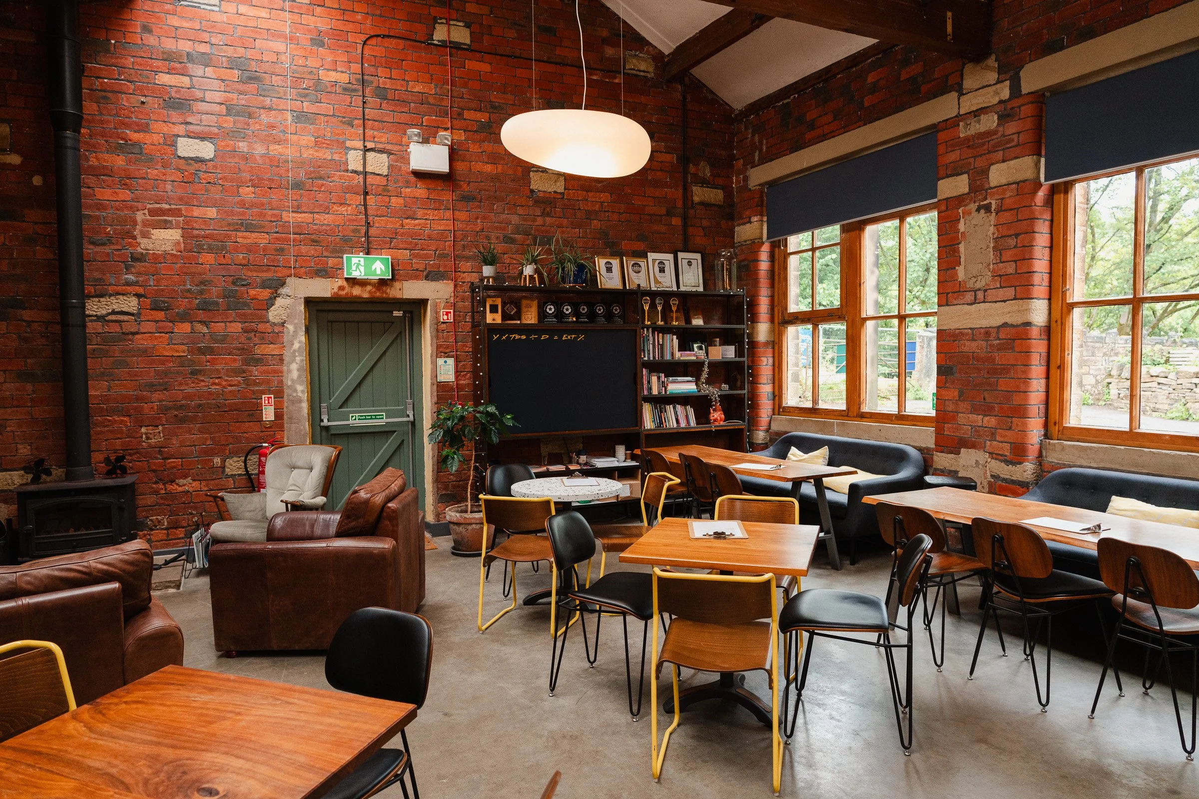 Interior of a communal space with exposed red brick walls, large windows with wooden frames, and a mix of wooden and upholstered chairs and tables. There's a black bookshelf with books and framed awards or certificates, a black chalkboard, plants on 