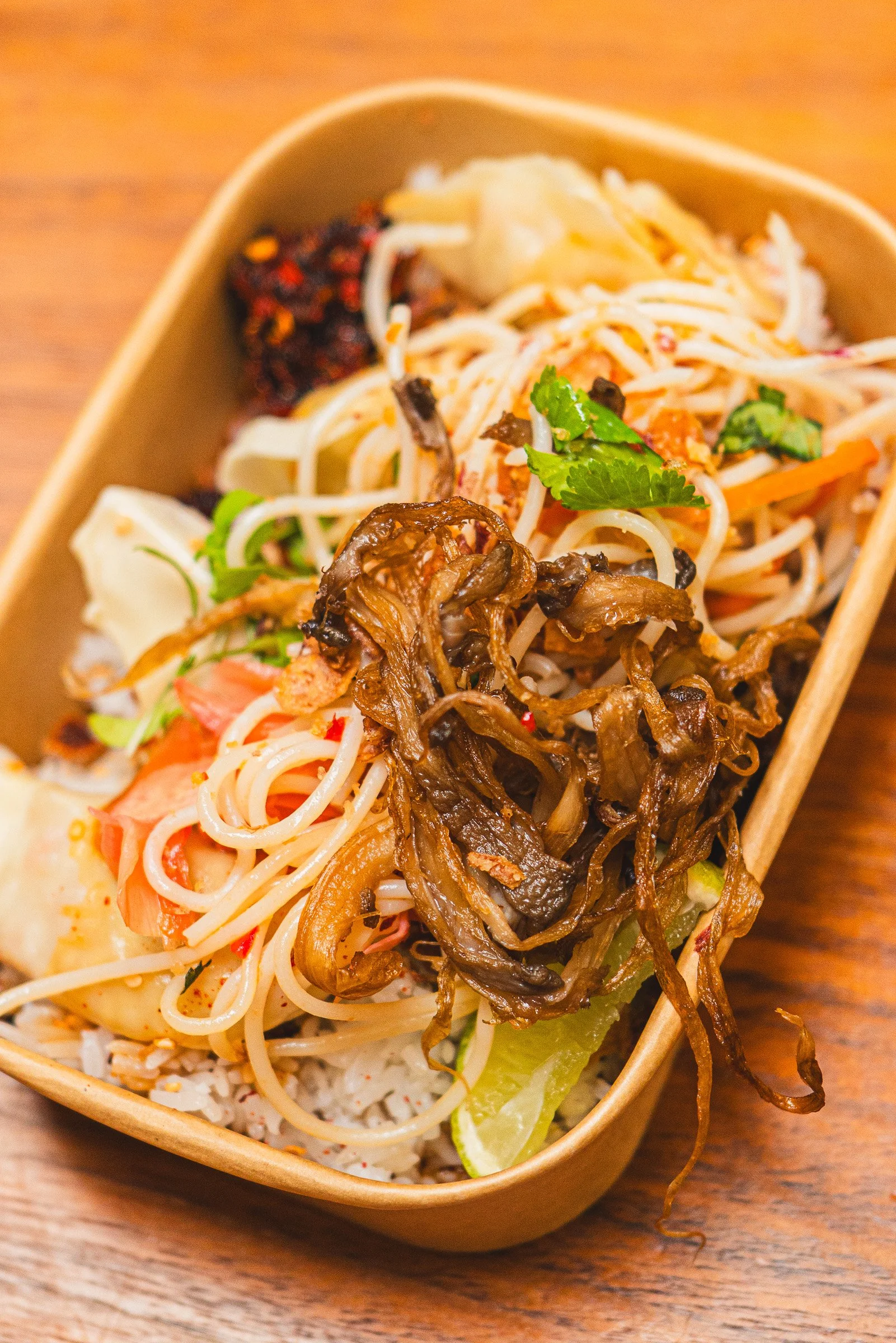 Close-up of a Thai noodle dish, herbs, and grilled vegetables, in a beige container on a wooden surface.