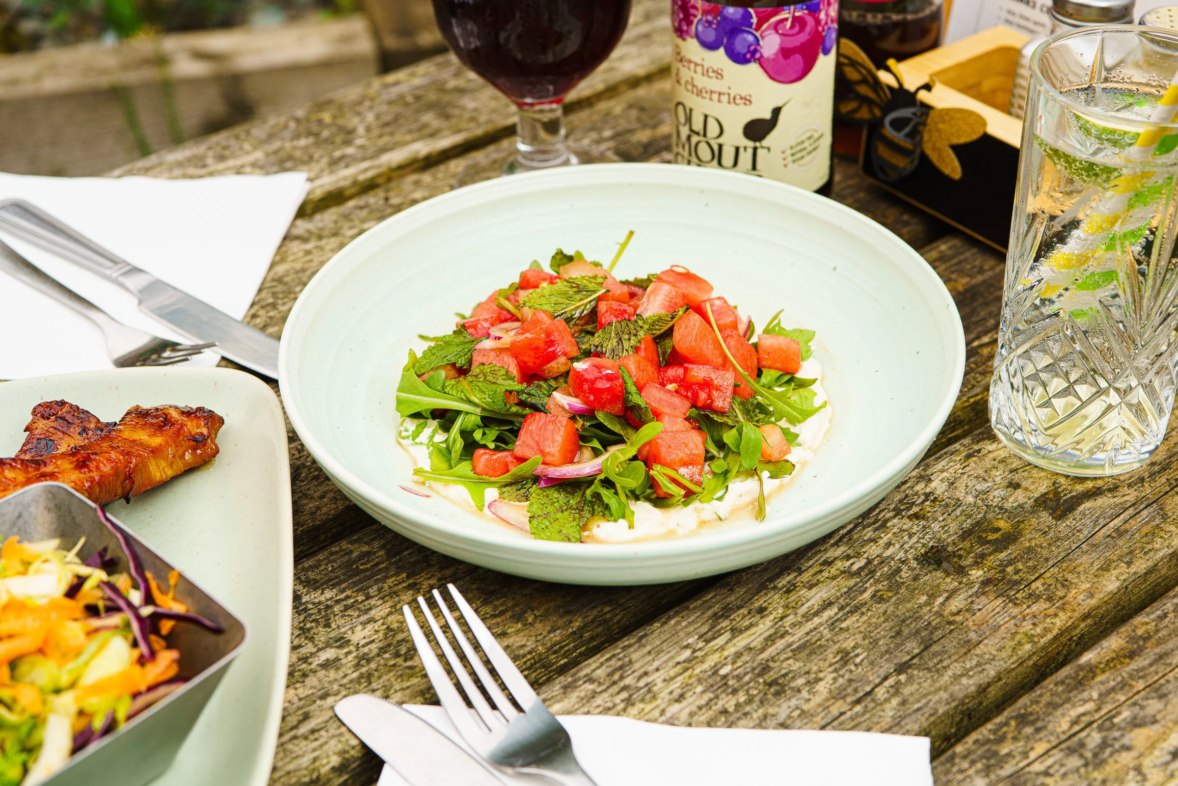 A bowl of salad with chopped watermelon, greens, and herbs on a rustic wooden table, with drinks and food in the background.