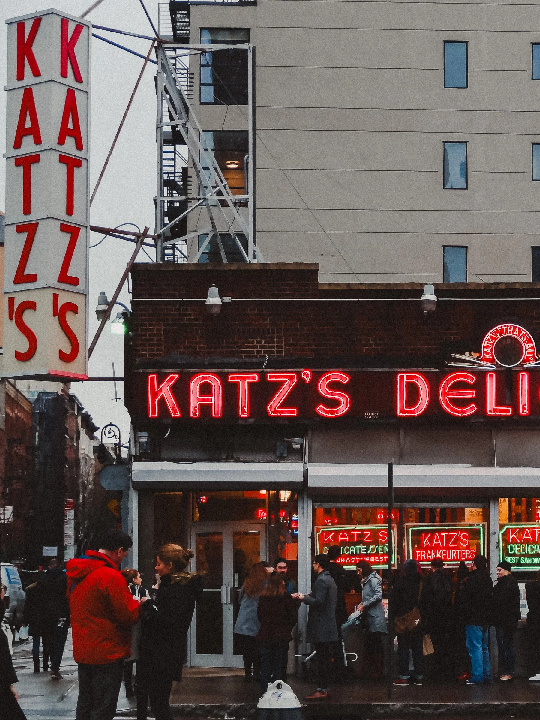 Crowd of people outside Katz's Deli in New York City with neon signs and a tall building in the background.