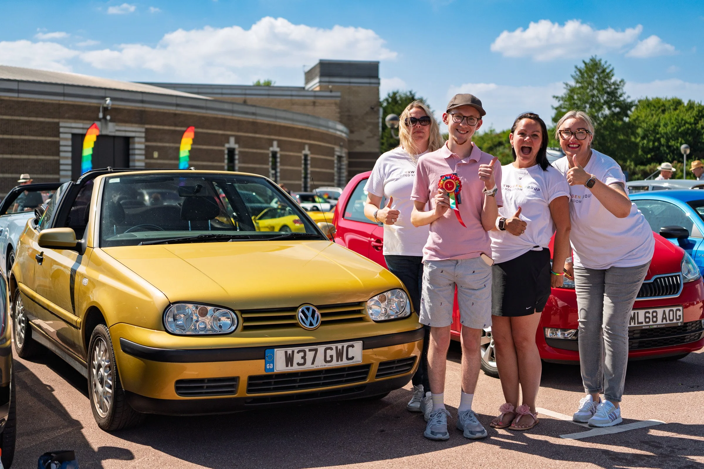 Group of four women and one man standing next to a yellow Volkswagen car at an outdoor event, with rainbow flags and other cars visible in the background on a sunny day.
