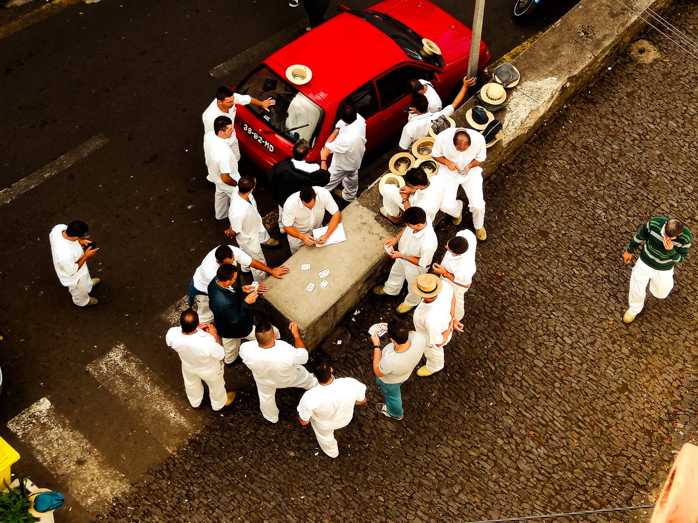 Group of people dressed in white clothing gathered on a cobblestone street around a parked red car, some appear to be engaged in a card game.