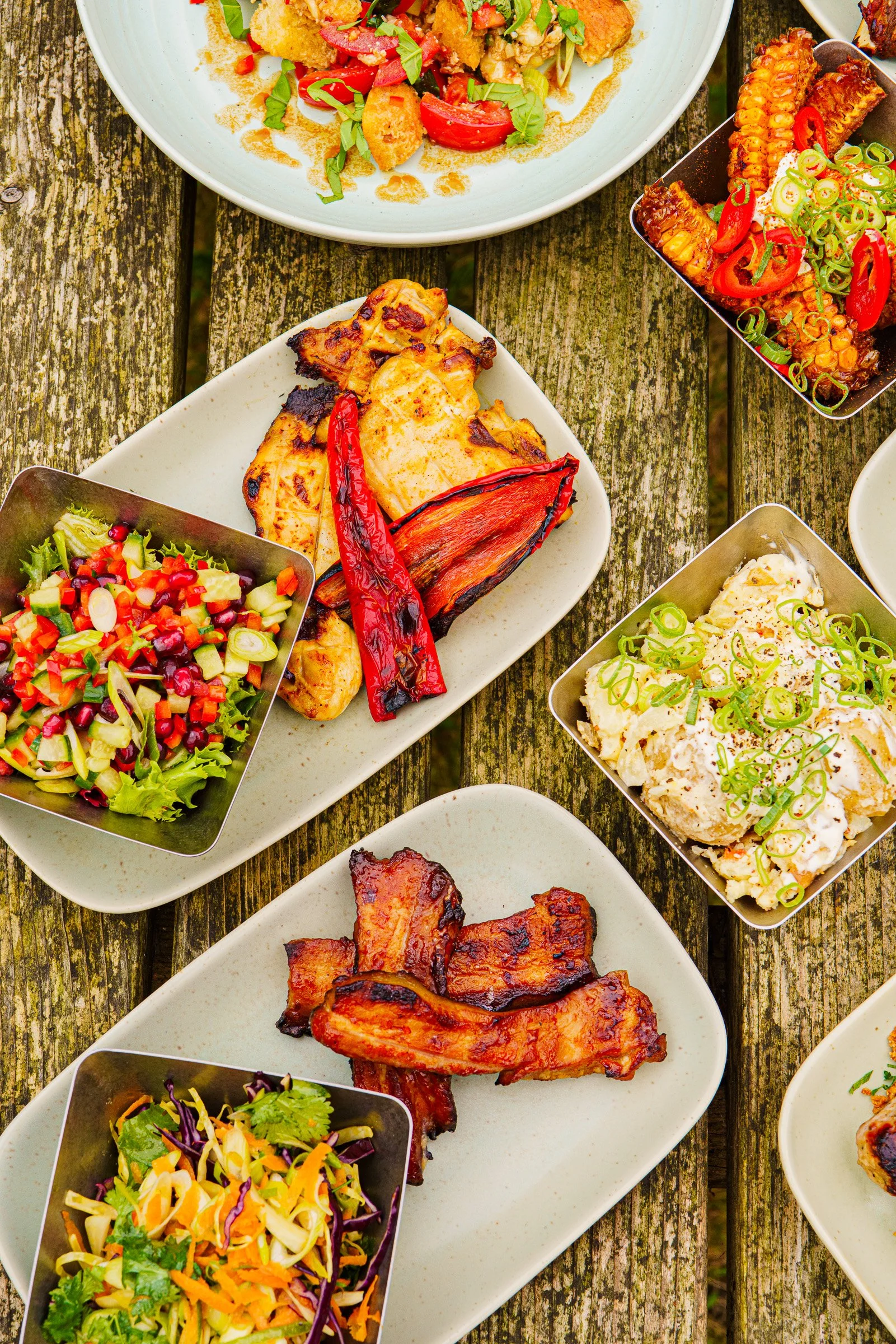 Various dishes including grilled chicken, salads, corn skewers, and barbecue ribs arranged on white and metal plates on a wooden table.