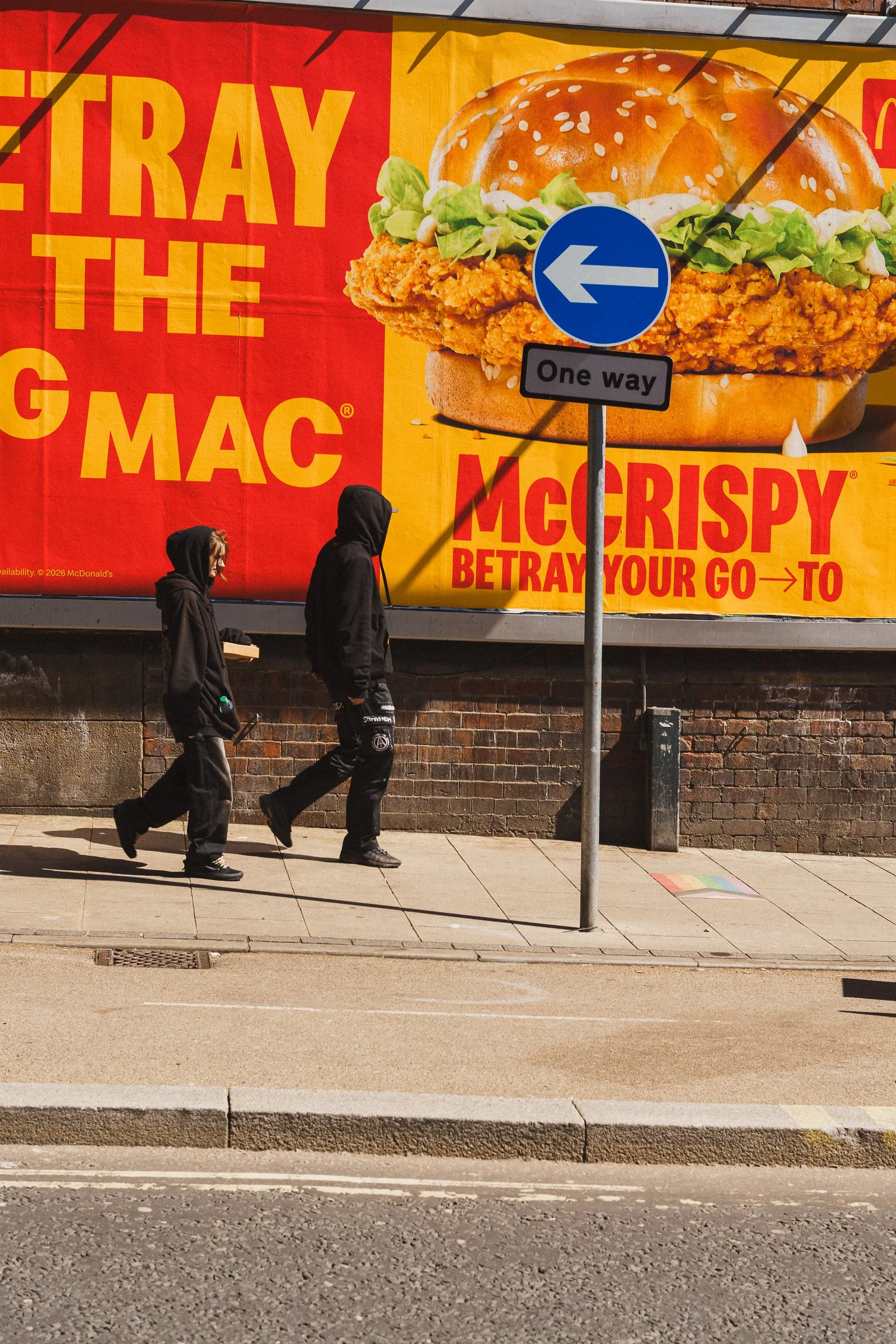 Two people walking on the sidewalk in front of a large McDonald's billboard advertisement. The billboard features a large image of a chicken sandwich. There is a one way street sign pointing the opposite direction to the way the two people are headed