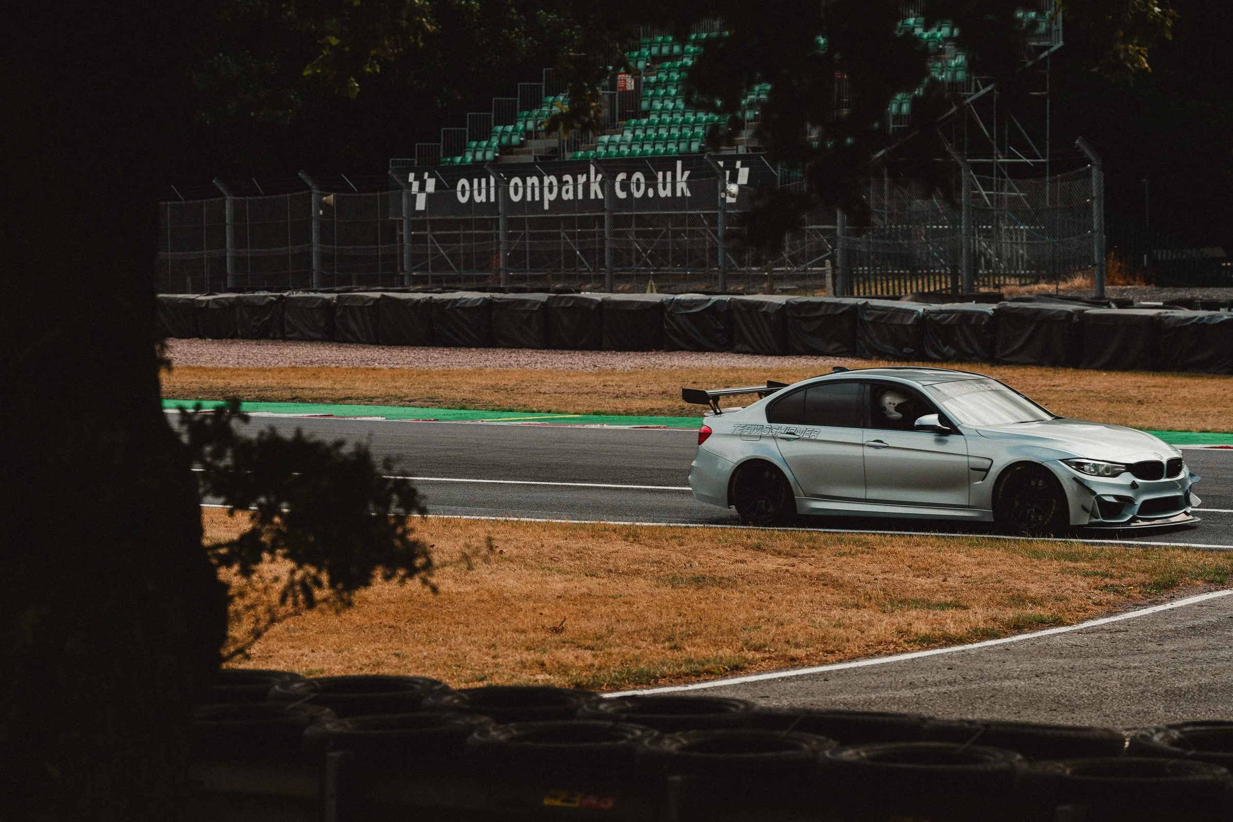 A silver sports car at Oulton Park, with green and black racing barriers behind it and green stadium seats in the background. The photo is taken from behind a tree creating negative space within the image.