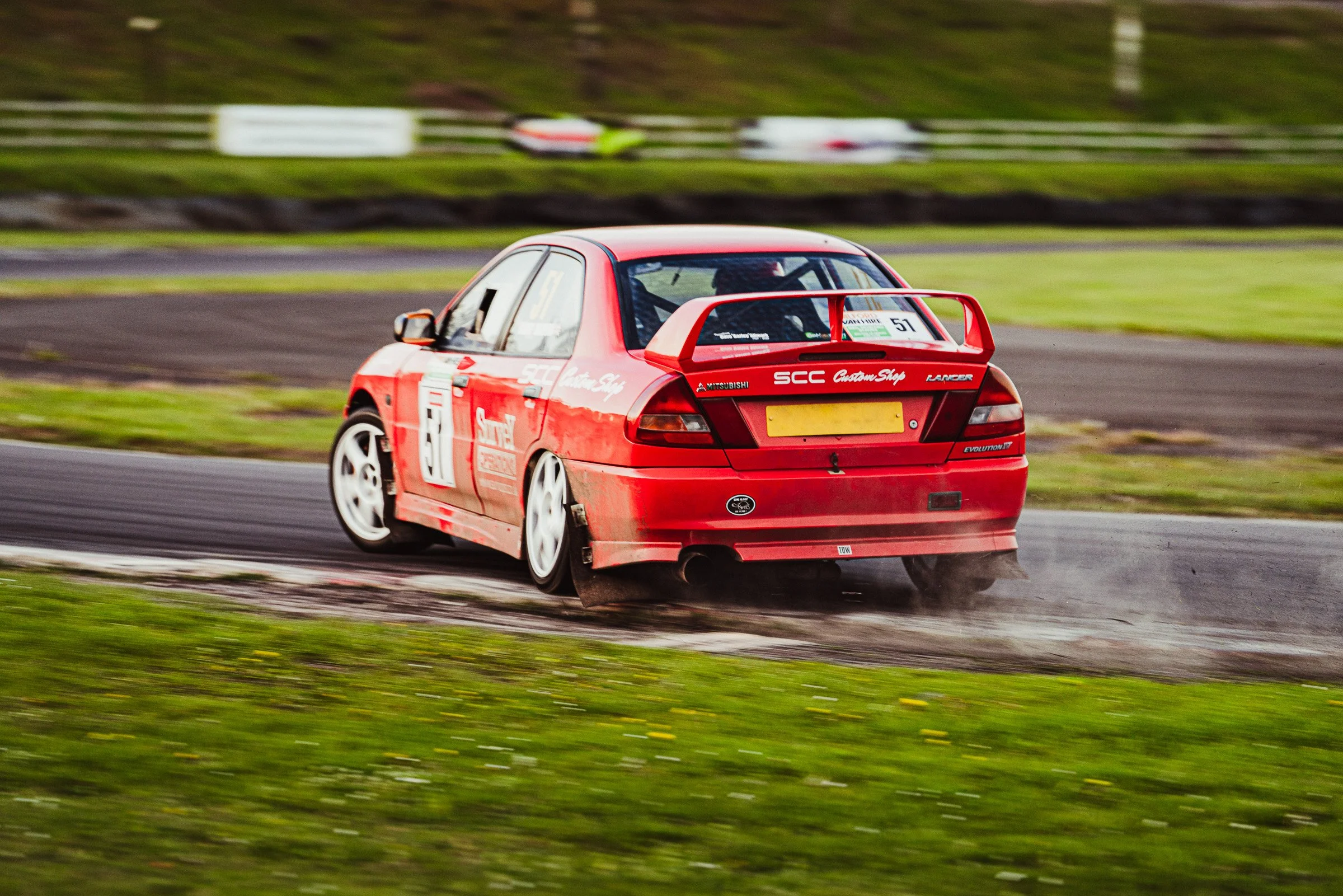 A red Mitsubishi race car is dropping a wheel on the gravel at a race track surrounded by grass, with motion blur in the background indicating high speed.