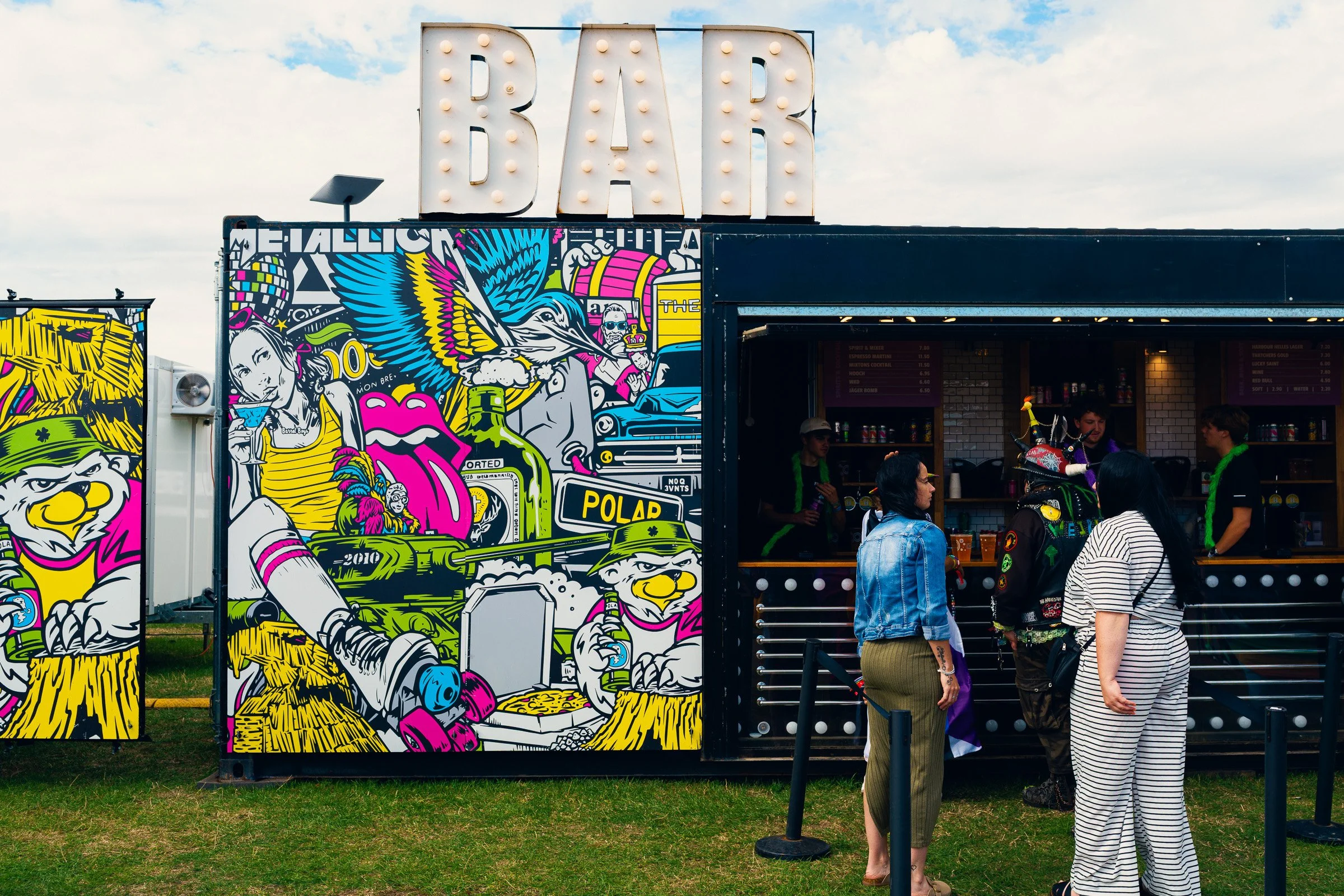 People standing outside a food stand with colorful graffiti art, including a dog, a woman holding a drink, and various abstract designs, under a cloudy sky.