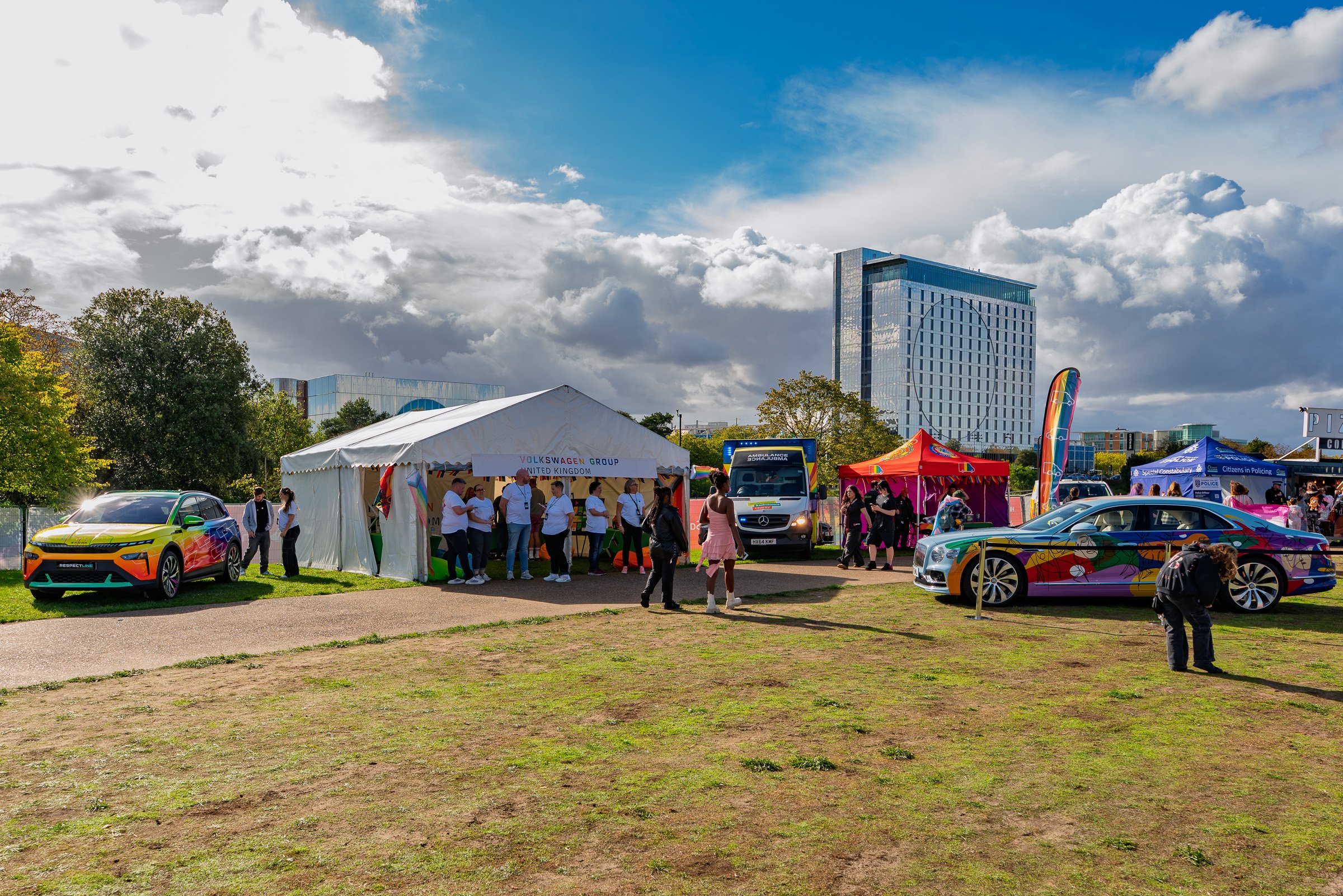 A vibrant outdoor event with people gathered around tents and parked cars, some with colorful designs, in a cityscape with modern high-rise buildings and a partly cloudy sky.