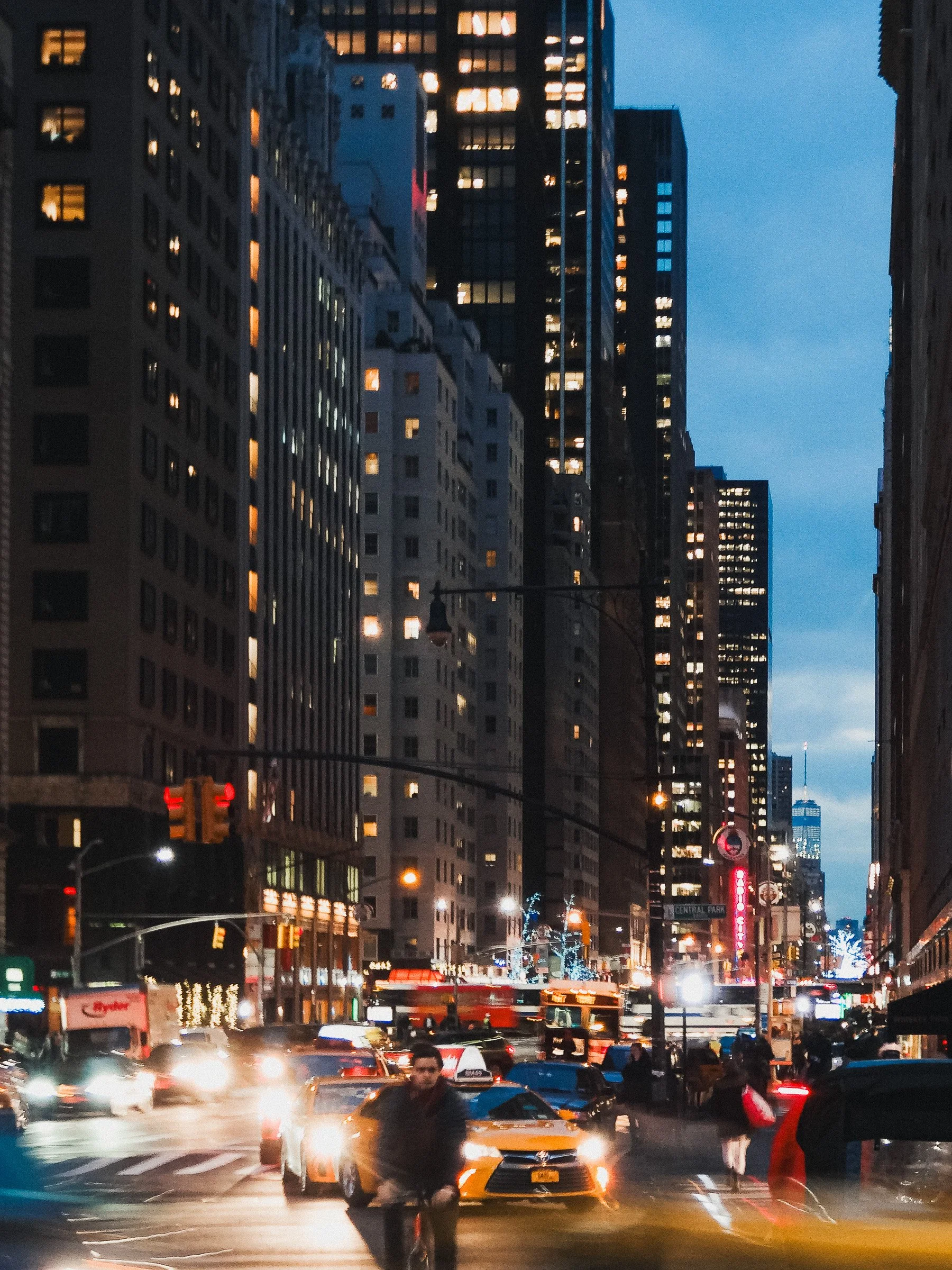 Night time New York city street scene with tall buildings, some with lights on. Traffic and pedestrians are visible, with cars and buses on the road. People cross the street in the foreground, and city lights illuminate the scene.