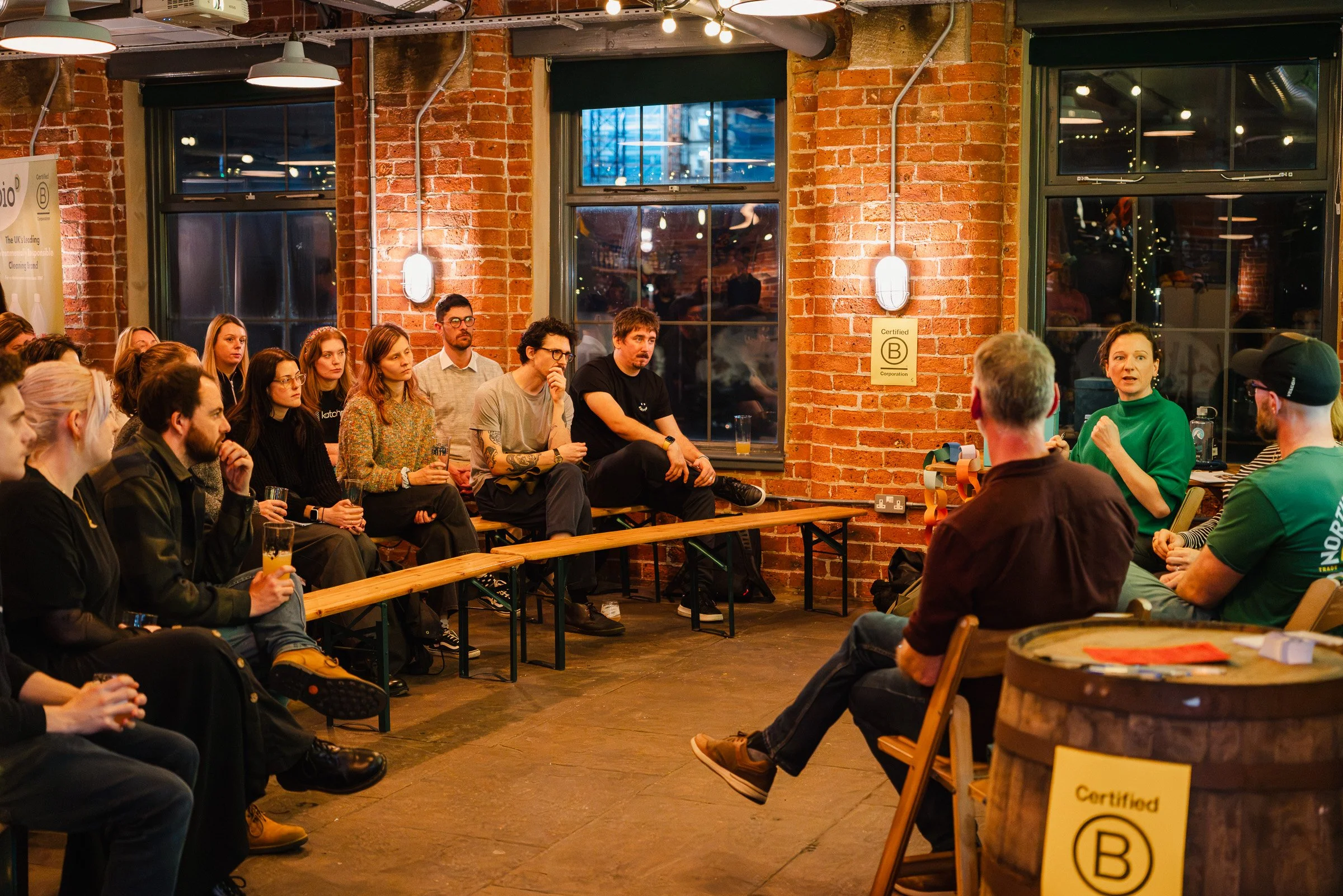 A group of people sitting in an industrial-style venue with exposed brick walls and large windows, attending a presentation or discussion led by a woman in a green shirt.