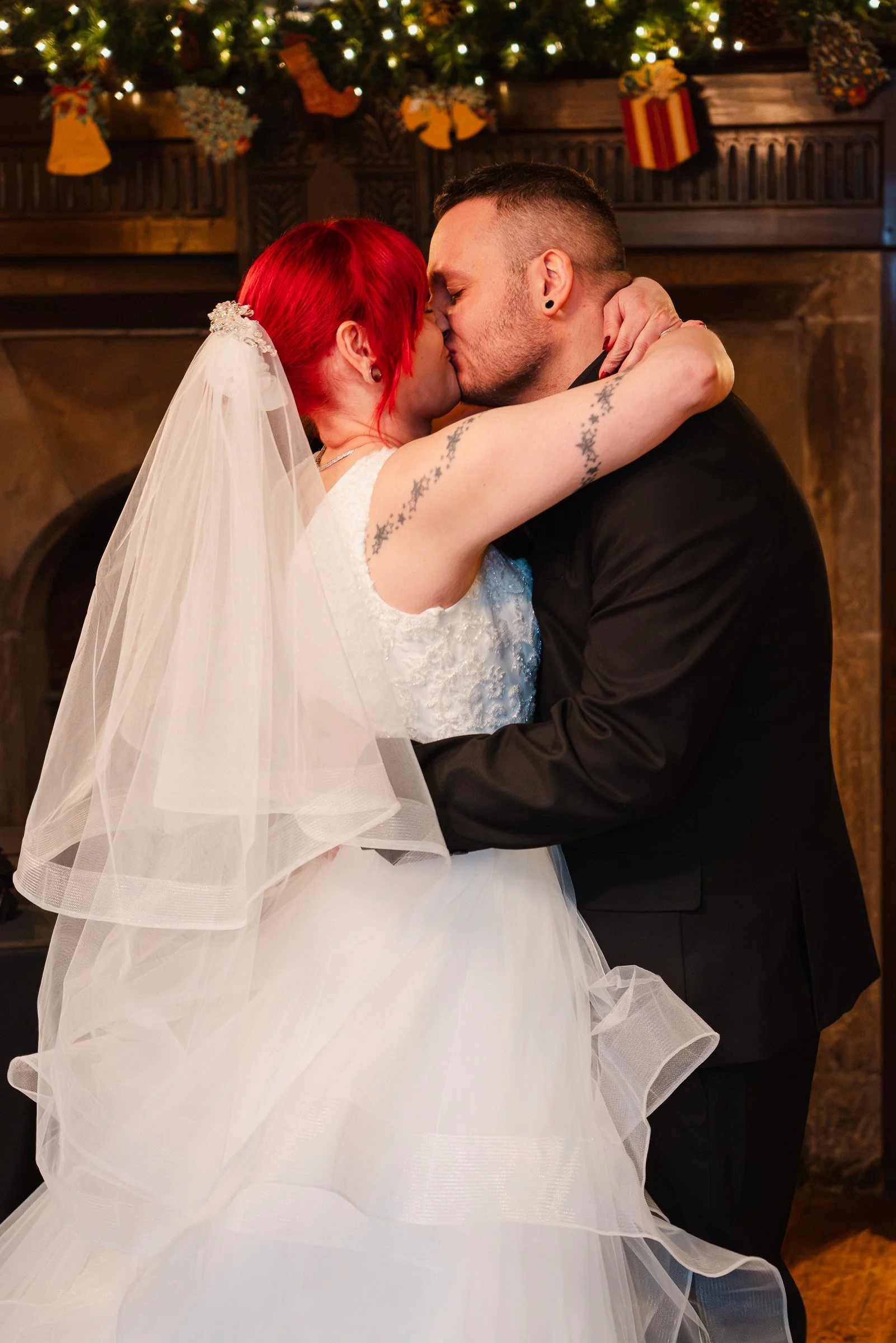 A bride and groom are kissing during a wedding ceremony in a cozy, decorated indoor setting with Christmas stockings and fairy lights in the background.