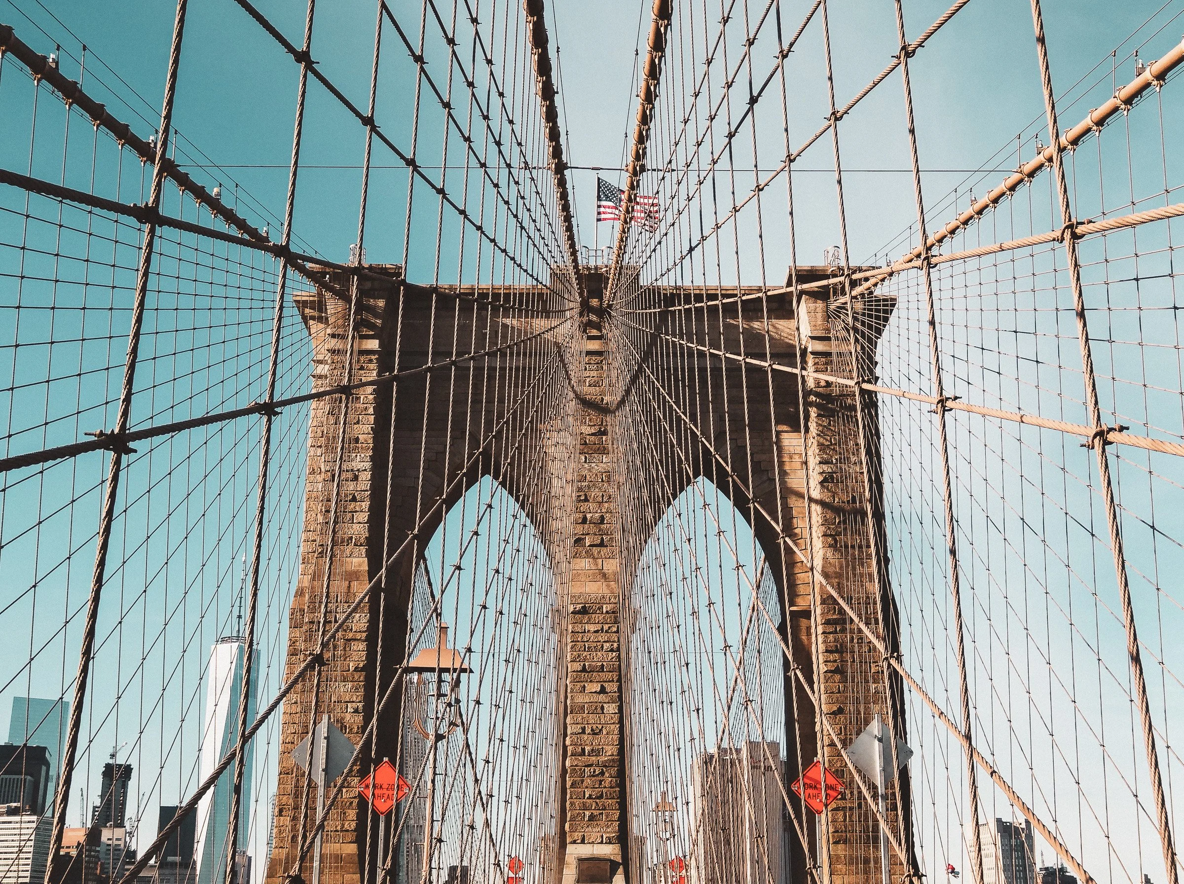 View looking up at the Brooklyn Bridge with its stone towers and crisscrossing steel cables, American flags atop the bridge, blue sky in background, and city buildings in the distance.