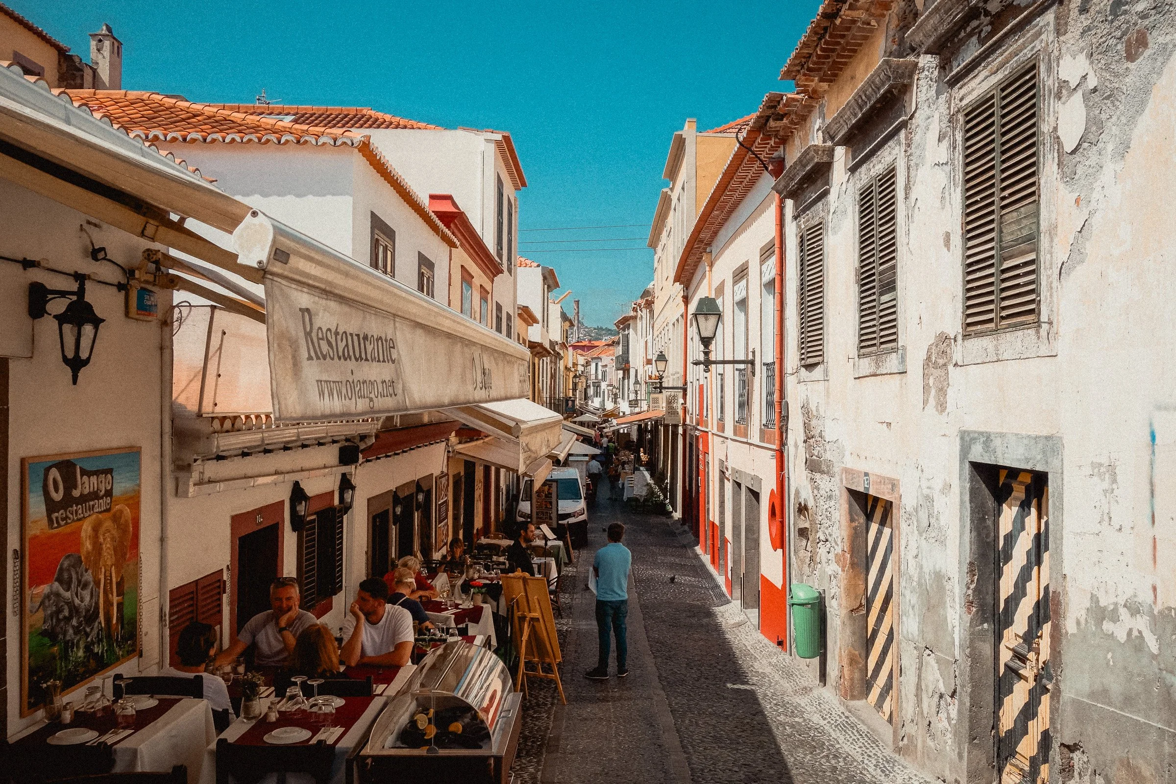 A lively street scene with outdoor dining, people socializing, and historic buildings with colorful facades, red-tiled roofs, and old shutters under a bright blue sky.