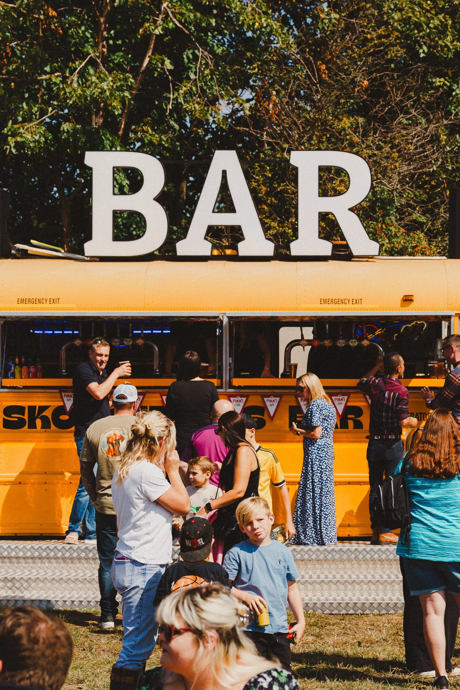 A yellow school bus with large white letters spelling 'BAR' on top, serving drinks to a crowd of people at an outdoor event.