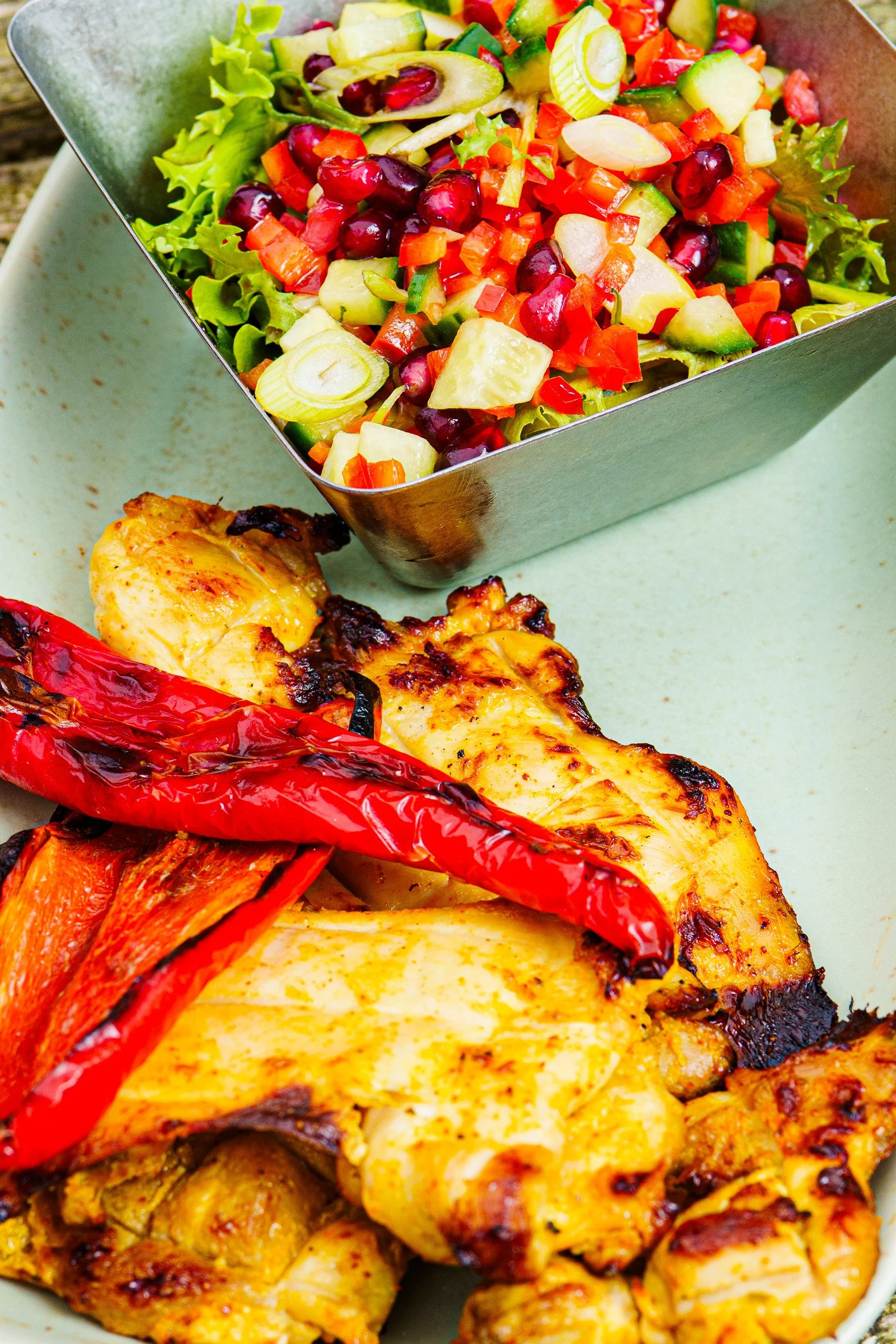 Close-up of a metal bowl filled with a colorful chopped vegetable salad containing cucumbers, cherry tomatoes, red bell peppers, green onions, and red grapes, placed on a light-colored surface next to grilled chicken and roasted red peppers.