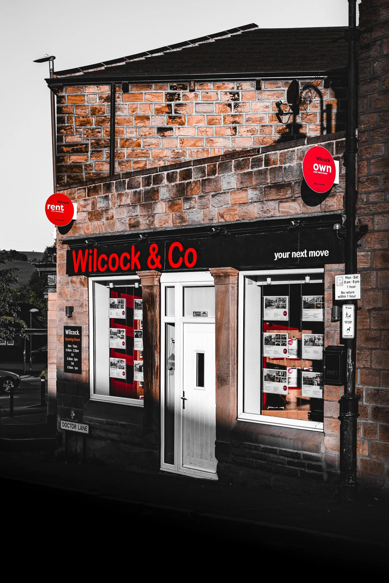 A brick building with signs for Wilcock & Co real estate agency, displaying property listings in the window. There are also signs for rent and own, with a black and red color theme, and an external street sign labeled Doctor Lane.