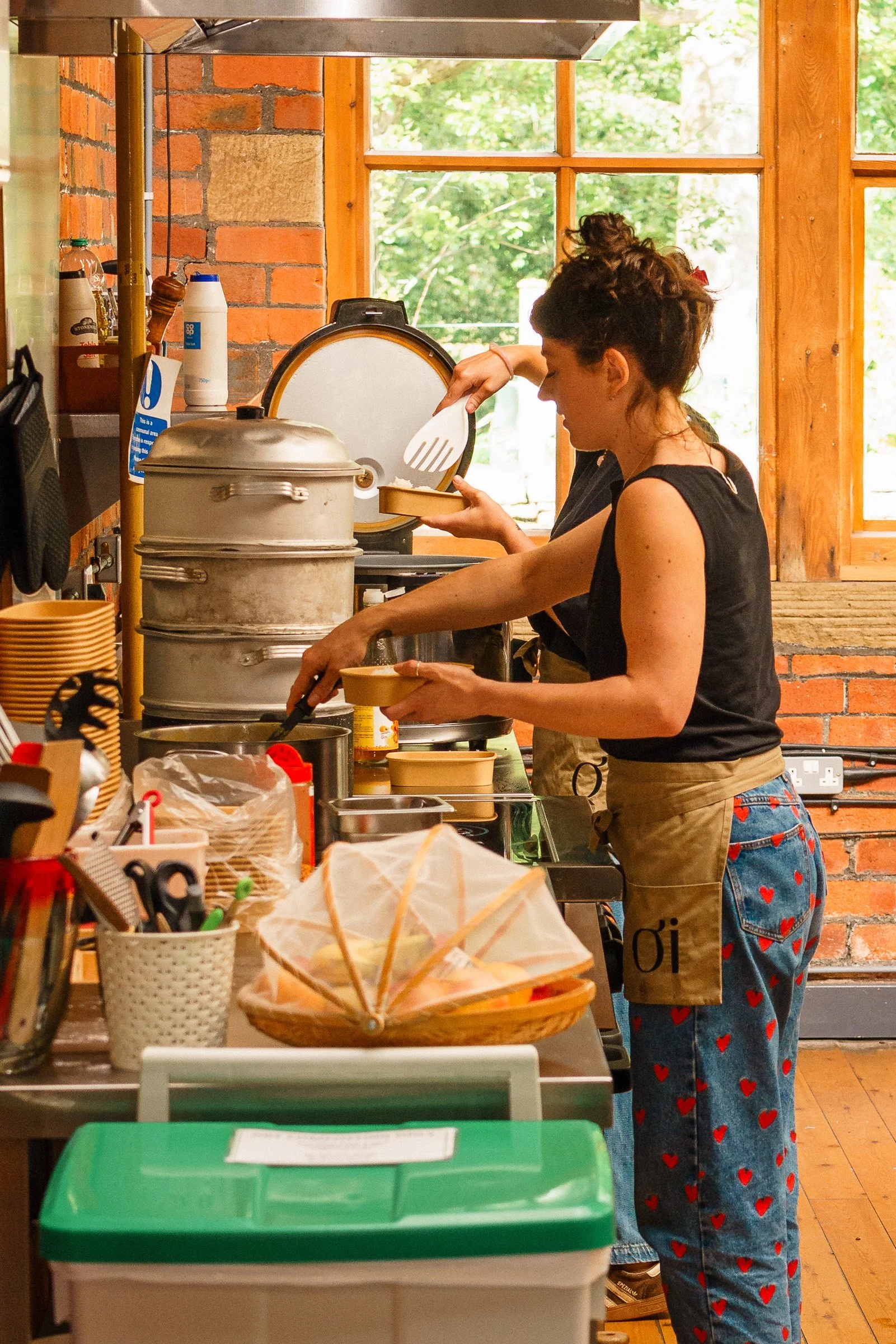 Two women preparing food in a rustic kitchen with brick walls and large windows. One woman is wearing a black top, an apron, and blue jeans with red heart patterns, while the other woman, partially visible, is wearing a dark shirt.