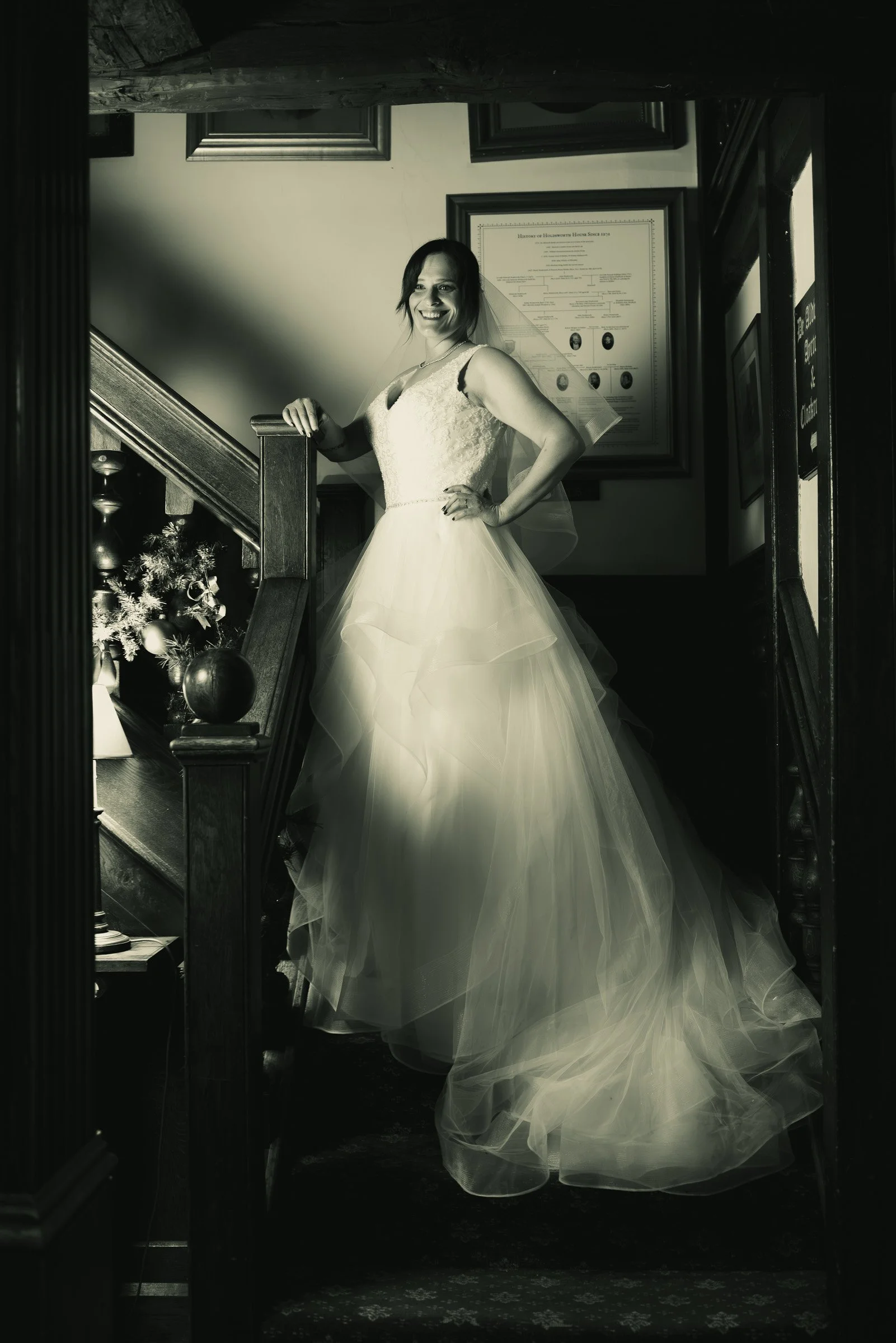 A woman in a wedding dress smiling, standing on a staircase in an indoor setting with framed documents on the wall behind her.