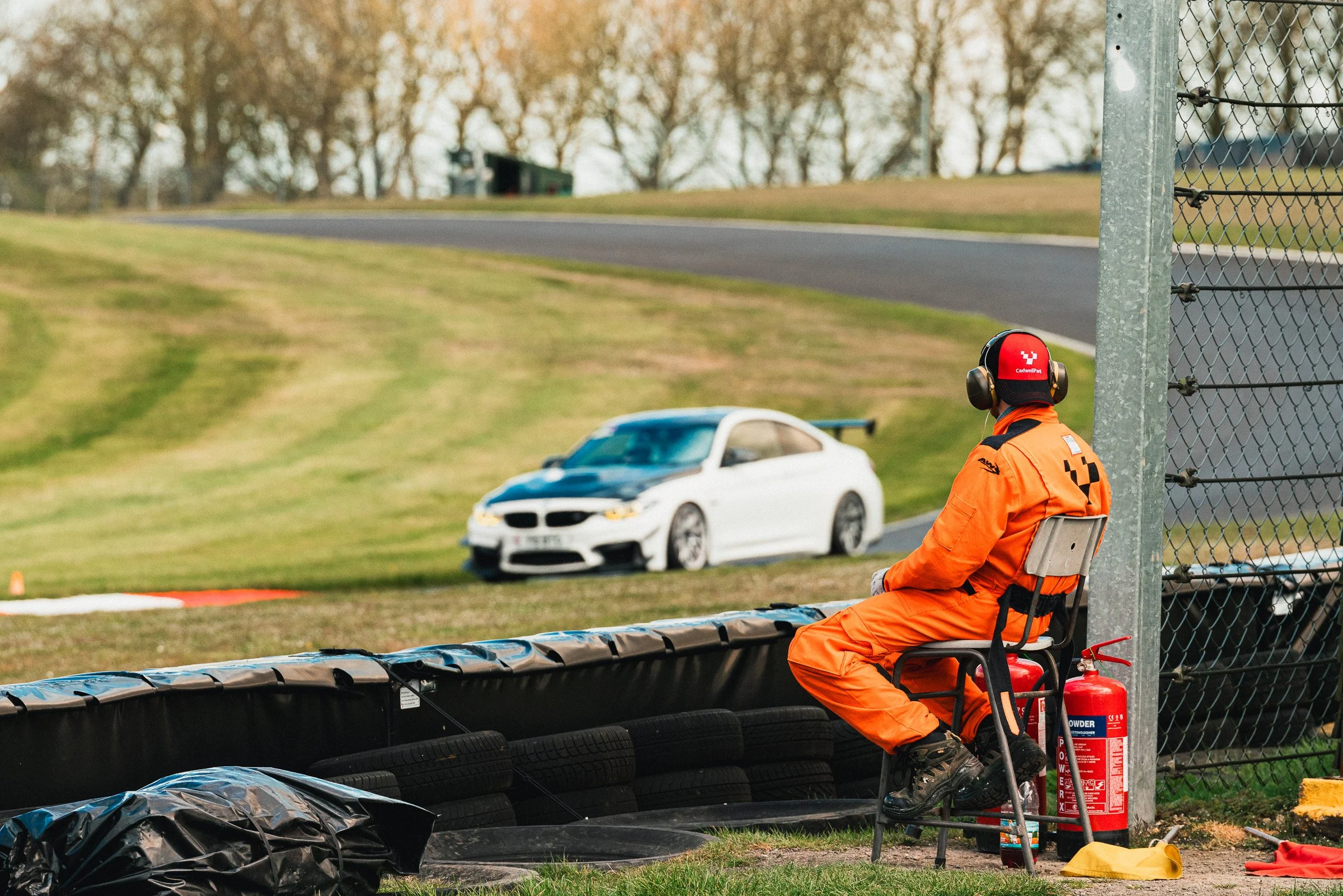 A race official sitting next to a tire barrier at a race track, wearing an orange safety suit and helmet, with a fire extinguisher and safety equipment nearby, overlooking a race car on the track.