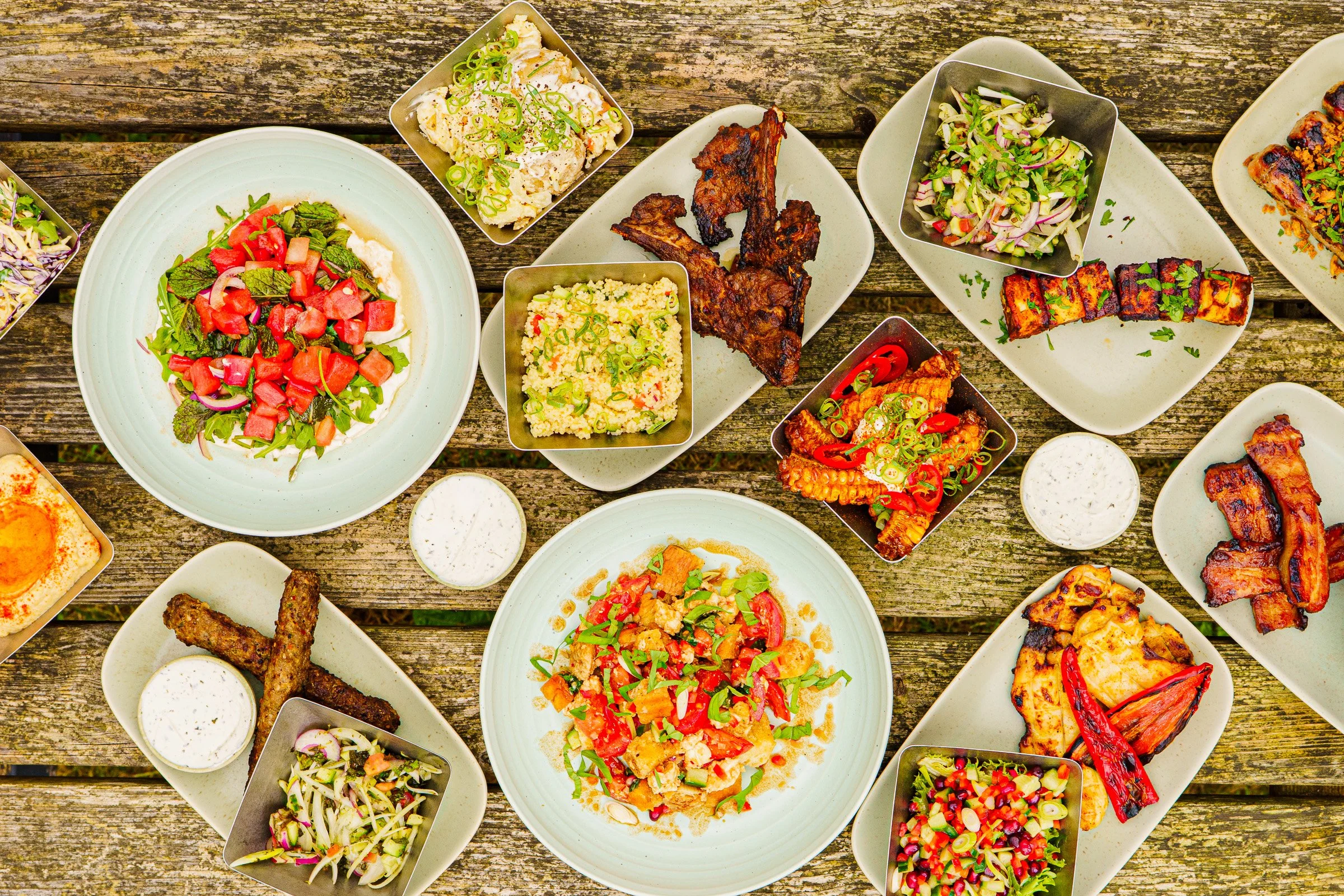 Assorted plates of barbecue dishes including grilled meats, salads, and side dishes on a rustic wooden table.