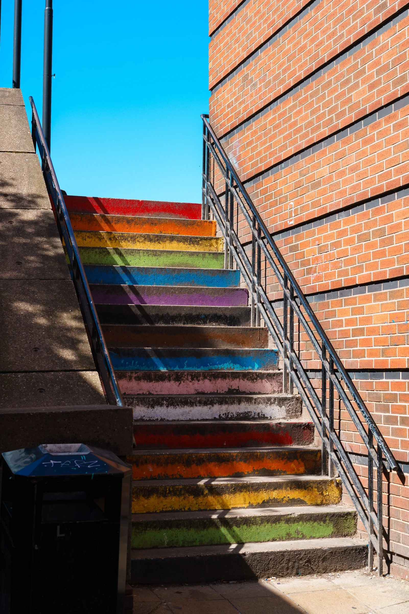 Outdoor staircase with rainbow-colored steps leading up to a brick building, with a bright blue sky above.