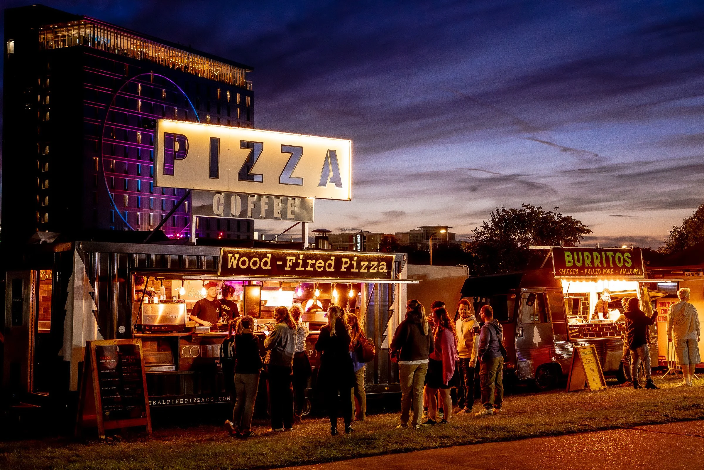 Night scene of a pizza food stand with neon lights and a large illuminated sign reading 'PIZZA.' There are people waiting and ordering, with food trucks nearby, and the sky is dark with some clouds.