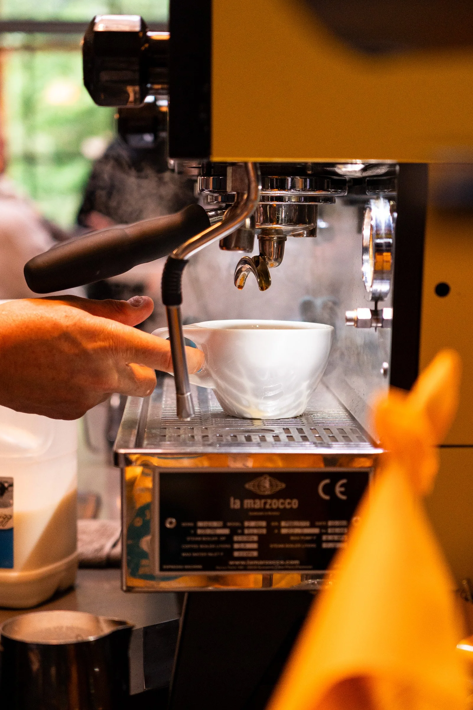 A person operating an espresso machine, preparing coffee in a white cup, with steam rising from the coffee.